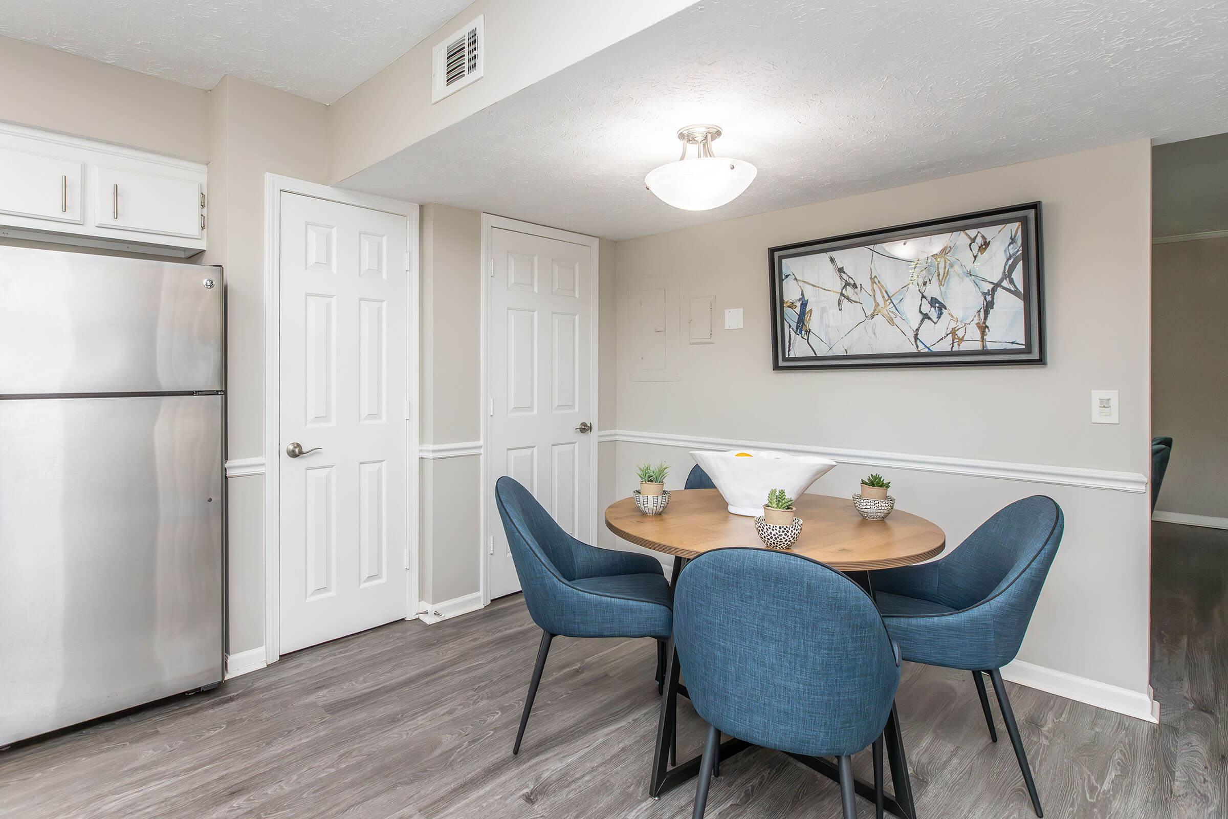 A cozy dining area featuring a round wooden table with four blue chairs. The table is adorned with small potted plants. A silver refrigerator is in the corner, and light-colored walls create a bright, inviting atmosphere. Above the table hangs a modern artwork in a black frame.