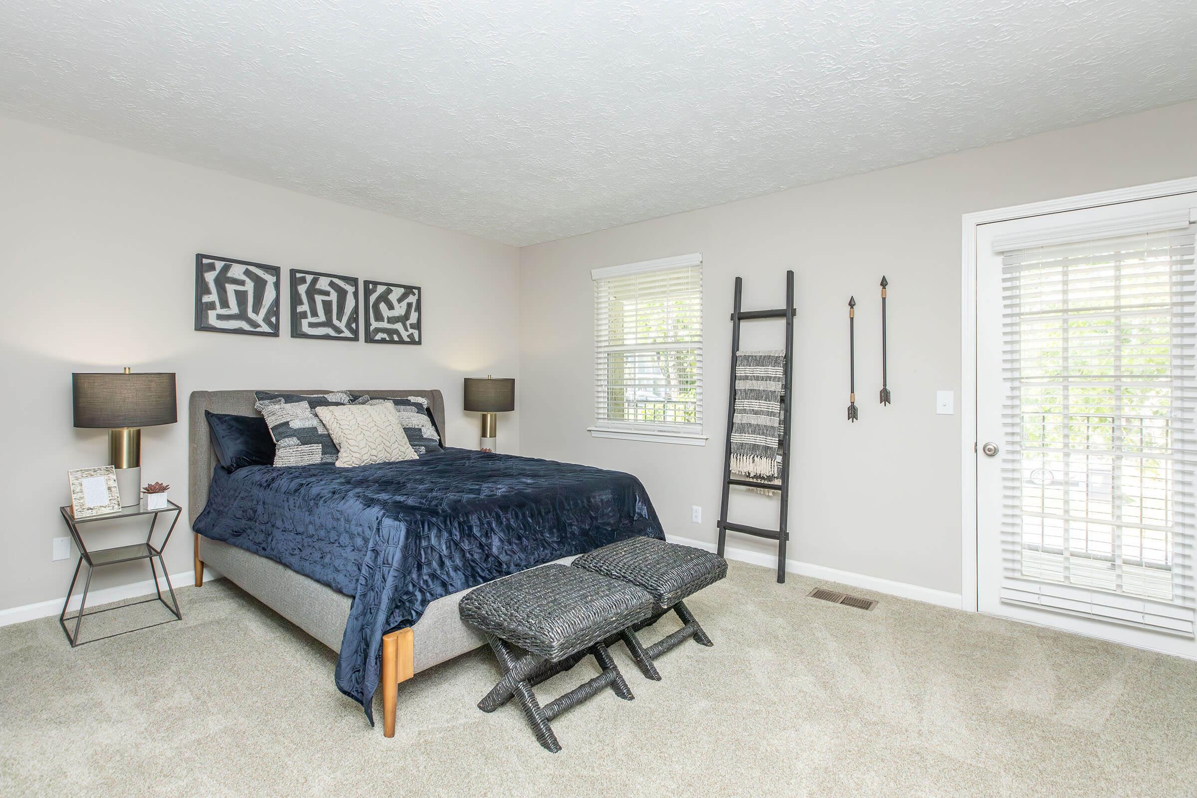 A cozy bedroom featuring a queen-sized bed with dark blue bedding, two bedside lamps, and abstract wall art. There is a decorative ladder against the wall, a patterned bench at the foot of the bed, and a sliding glass door leading to an outdoor area. Soft carpet flooring completes the inviting ambiance.