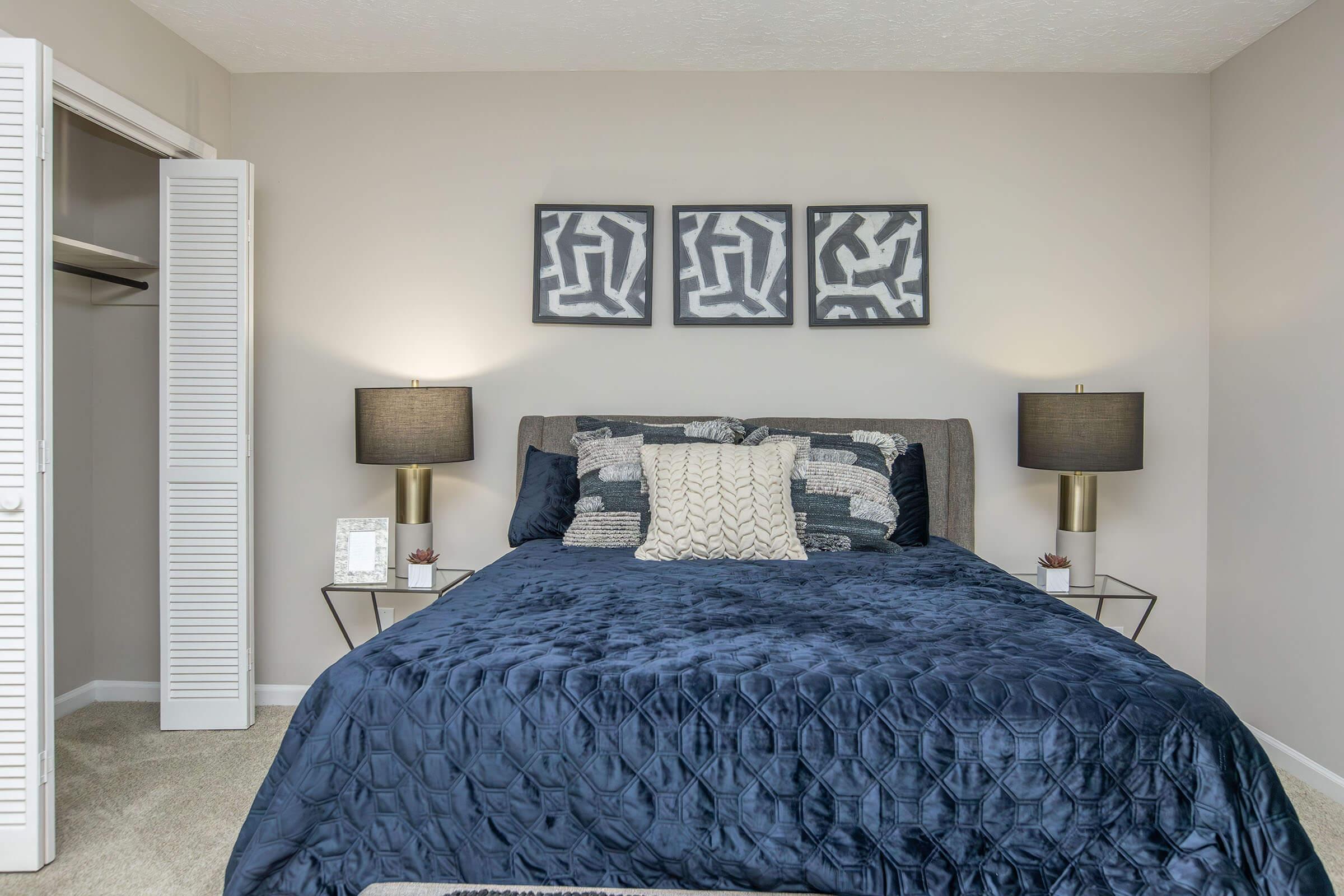 A cozy bedroom featuring a queen-sized bed with a navy blue quilt and multiple decorative pillows. On either side of the bed, there are stylish lamps on small end tables. Above the bed, there are three abstract framed artworks. A closet with white louvered doors is visible in the background. The walls are painted a soft beige.