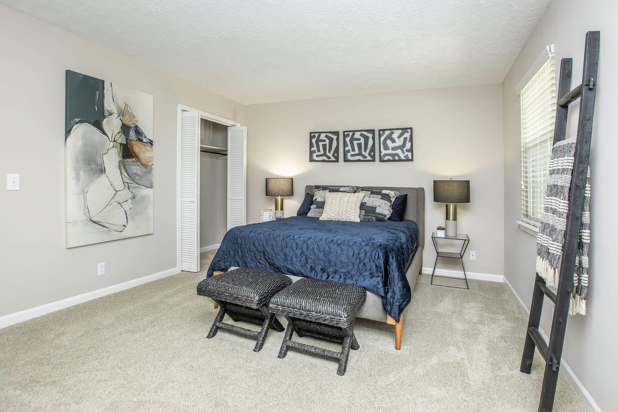 A modern bedroom featuring a neatly made bed with navy blue bedding, two decorative pillows, and two black woven stools at the foot. The walls are painted light gray with an abstract artwork above the bed and three framed prints. There's a closet and large windows allowing natural light to enter.