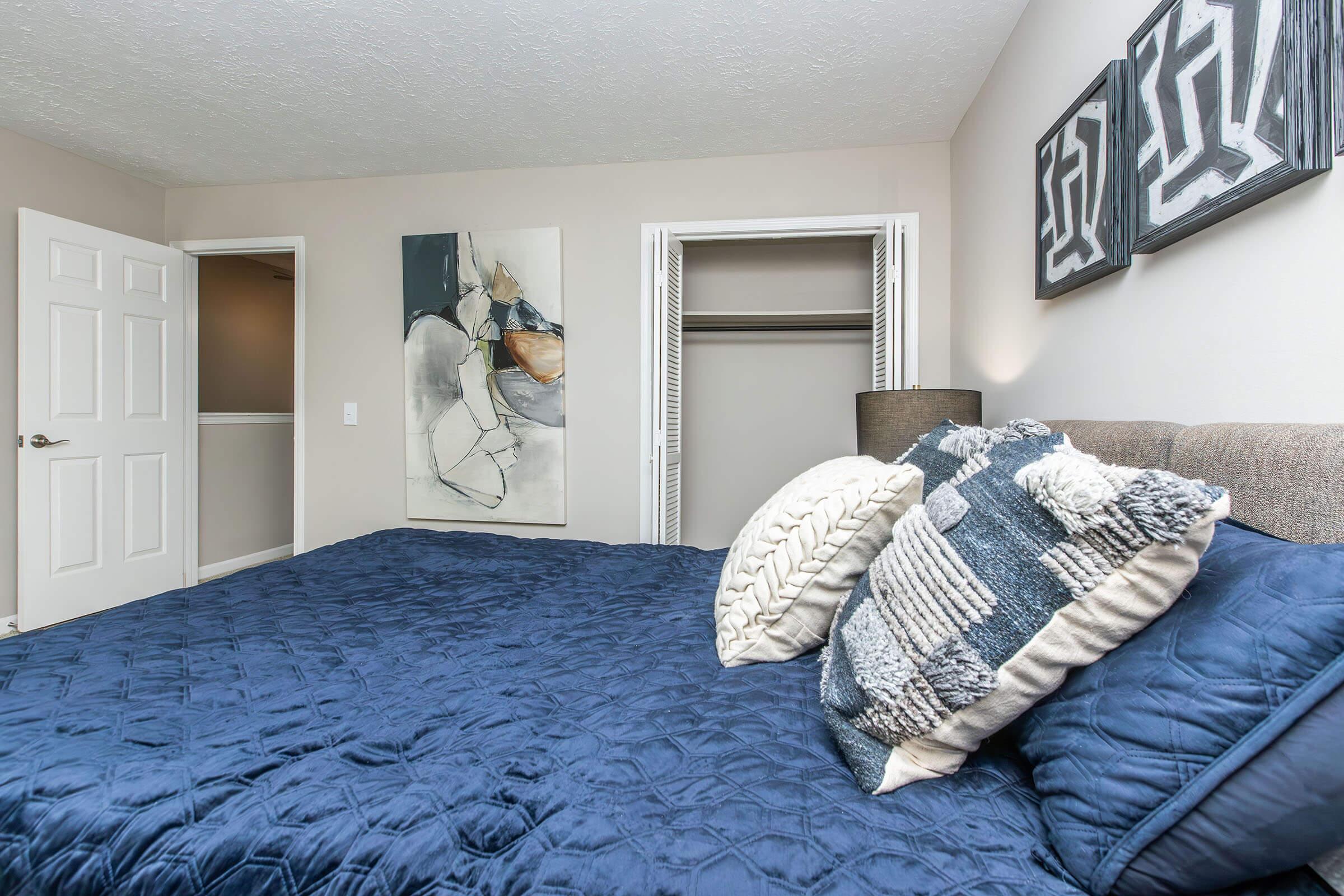 A cozy bedroom featuring a dark blue quilted bedspread with decorative pillows. The walls are painted in a neutral tone, and there are two abstract framed artworks above the bed. A closet door is slightly open in the background, and a doorway leads to another room. Soft lighting enhances the inviting atmosphere.