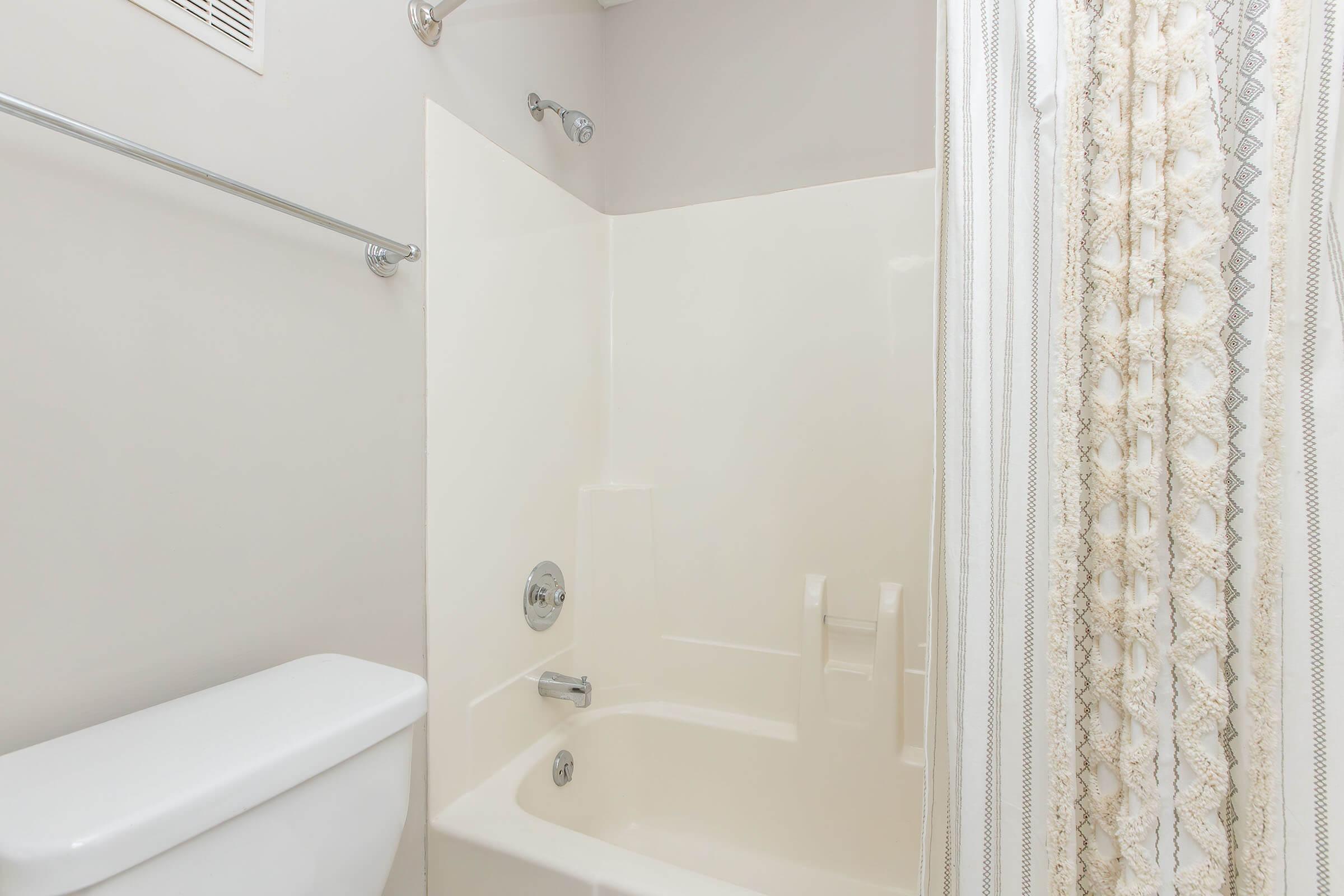 A clean bathroom with a white bathtub and shower combination. The walls are a light color, and there is a decorative shower curtain featuring a pattern. A towel bar is mounted on the wall, and a toilet is visible in the foreground. The space appears bright and well-maintained.