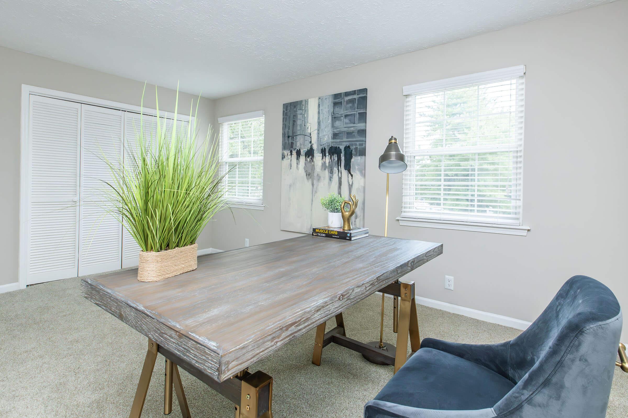 A modern home office featuring a large wooden desk with a decorative plant in a woven pot. To the side, there is a comfortable blue chair and a stylish lamp on the desk. The room is brightly lit with natural light coming through two windows, and a piece of abstract art hangs on the wall. Neutral-colored walls and carpet complete the setting.