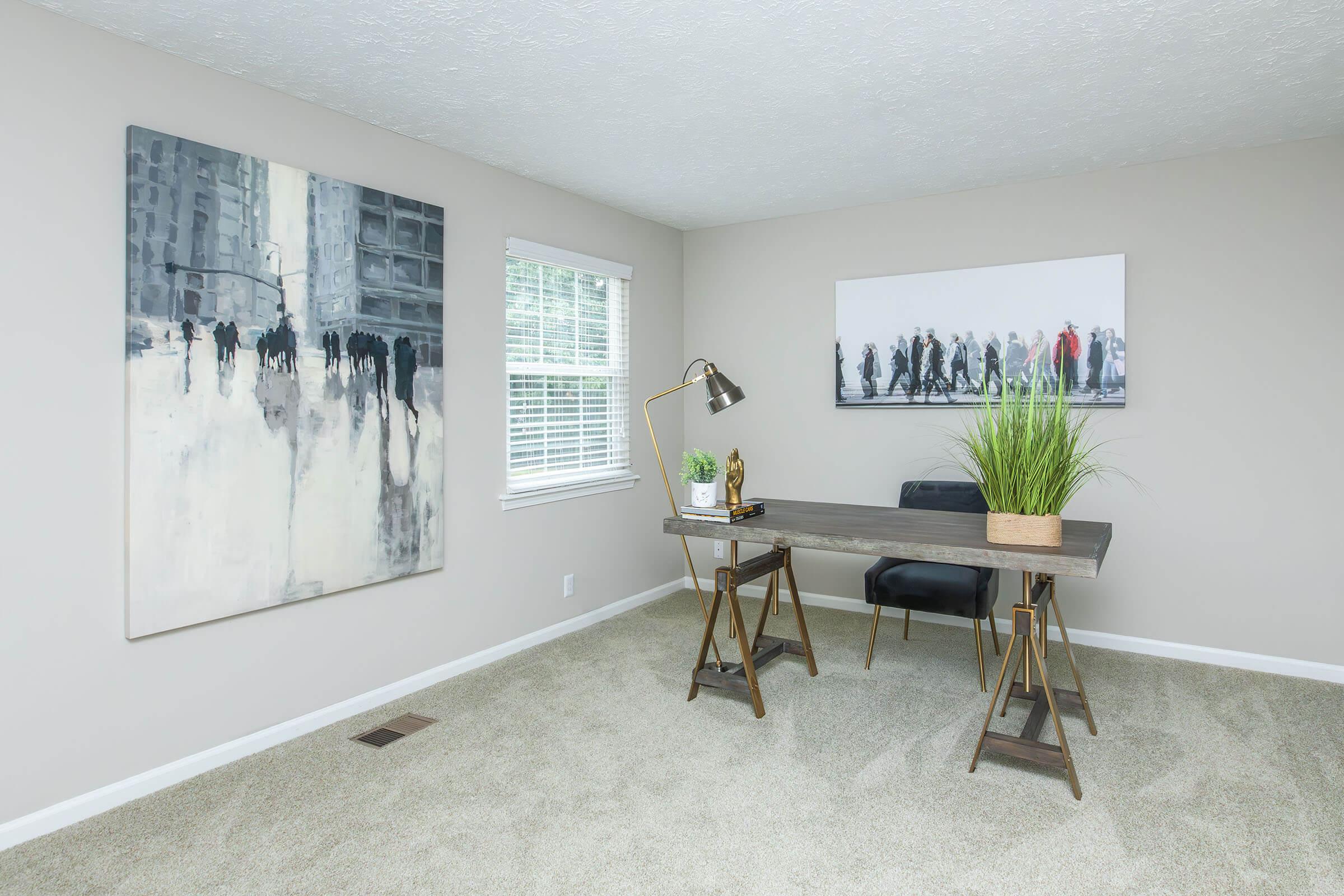 A modern, minimalistic home office featuring a wooden desk with two chairs, a table lamp, and a decorative plant. The room has light-colored walls and large windows allowing natural light. Two abstract paintings adorn the walls, adding a contemporary touch to the space. The carpeted floor enhances the cozy atmosphere.
