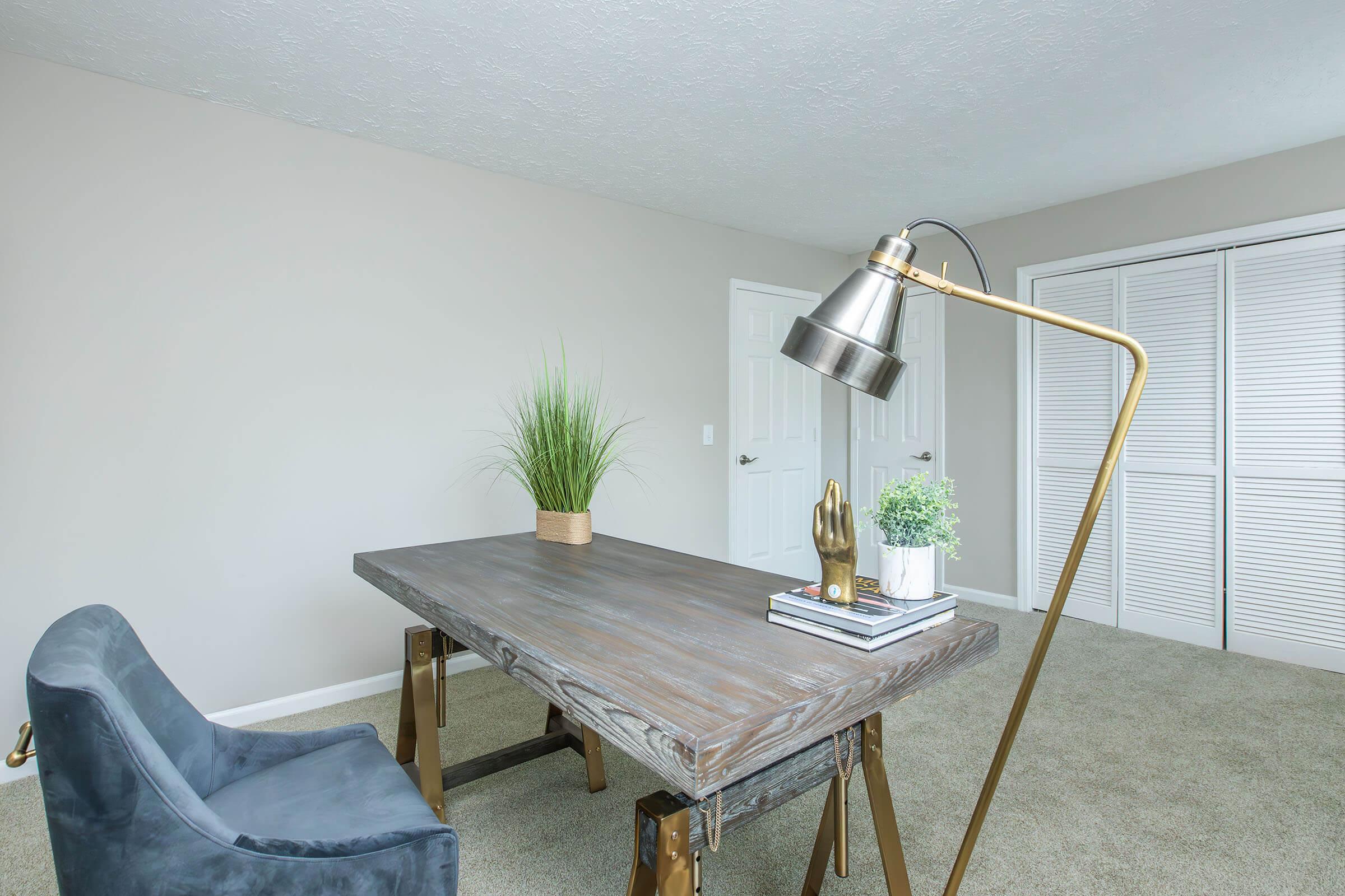 A modern office space featuring a wooden desk with a sleek design, a metallic desk lamp, a potted plant, decorative items on the desk, and a comfortable blue chair. The room has light, neutral-colored walls and a closet with sliding doors, creating a bright and inviting work environment.