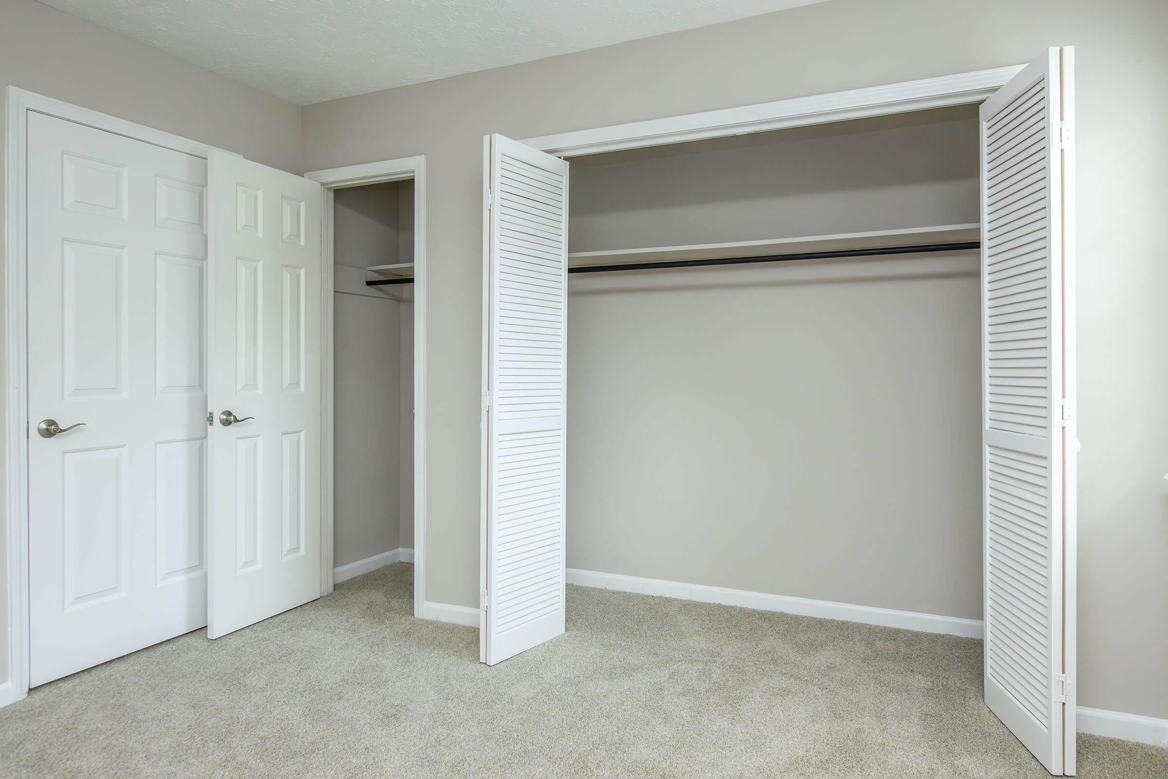 A spacious, empty room with light beige walls and carpet. There are two white doors open, revealing an empty closet with a shelf. The room is well-lit and uncluttered, creating a clean, minimalist aesthetic.