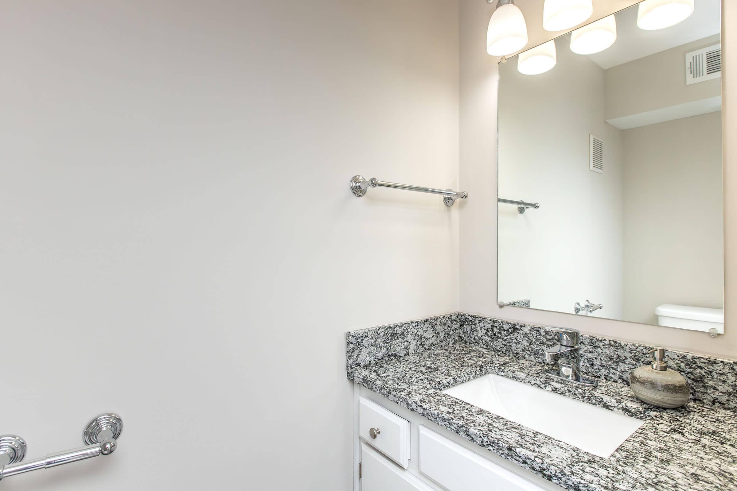 A modern bathroom featuring a granite countertop with a white sink and a decorative vase. Above the sink, there is a large mirror illuminated by three pendant lights. The walls are painted a neutral color, and a towel bar is mounted on one side.