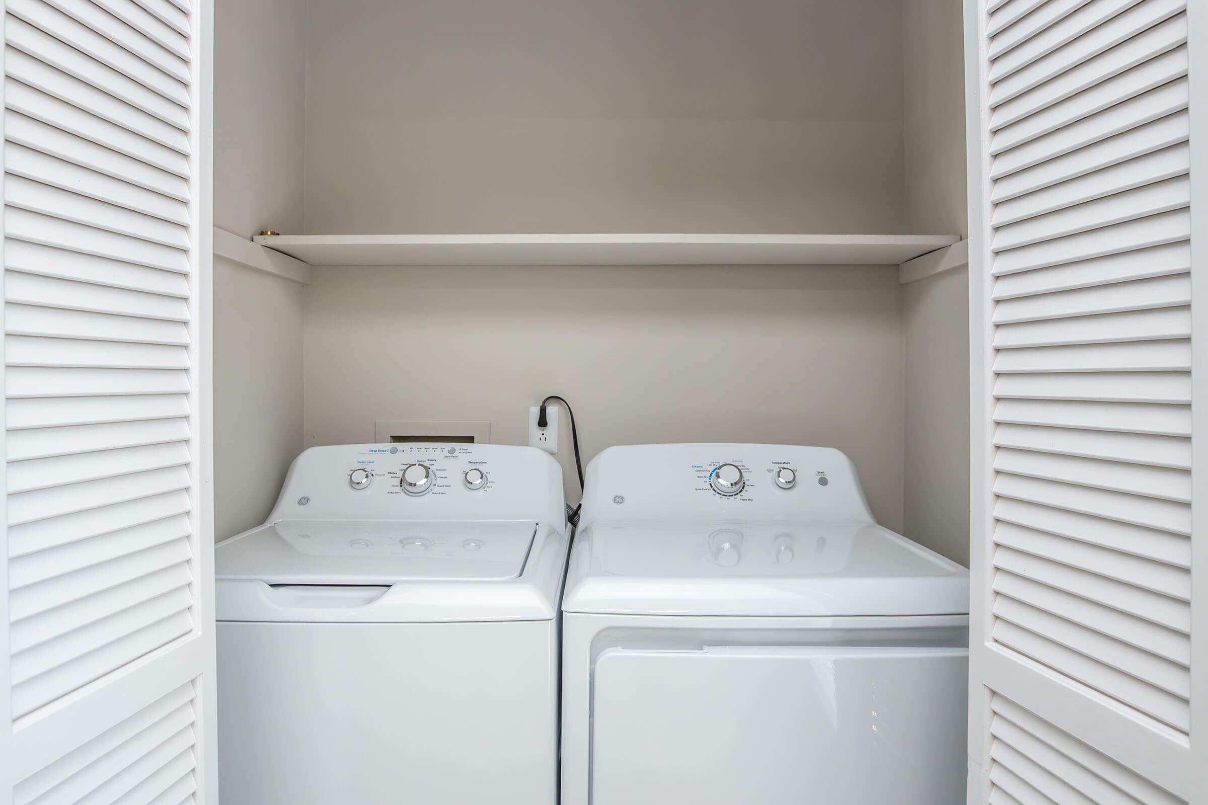 A compact laundry area featuring a white washing machine and a white dryer side by side. Above them, there is a shelf providing storage space. The space is enclosed by shuttered doors, creating a tidy and organized look.