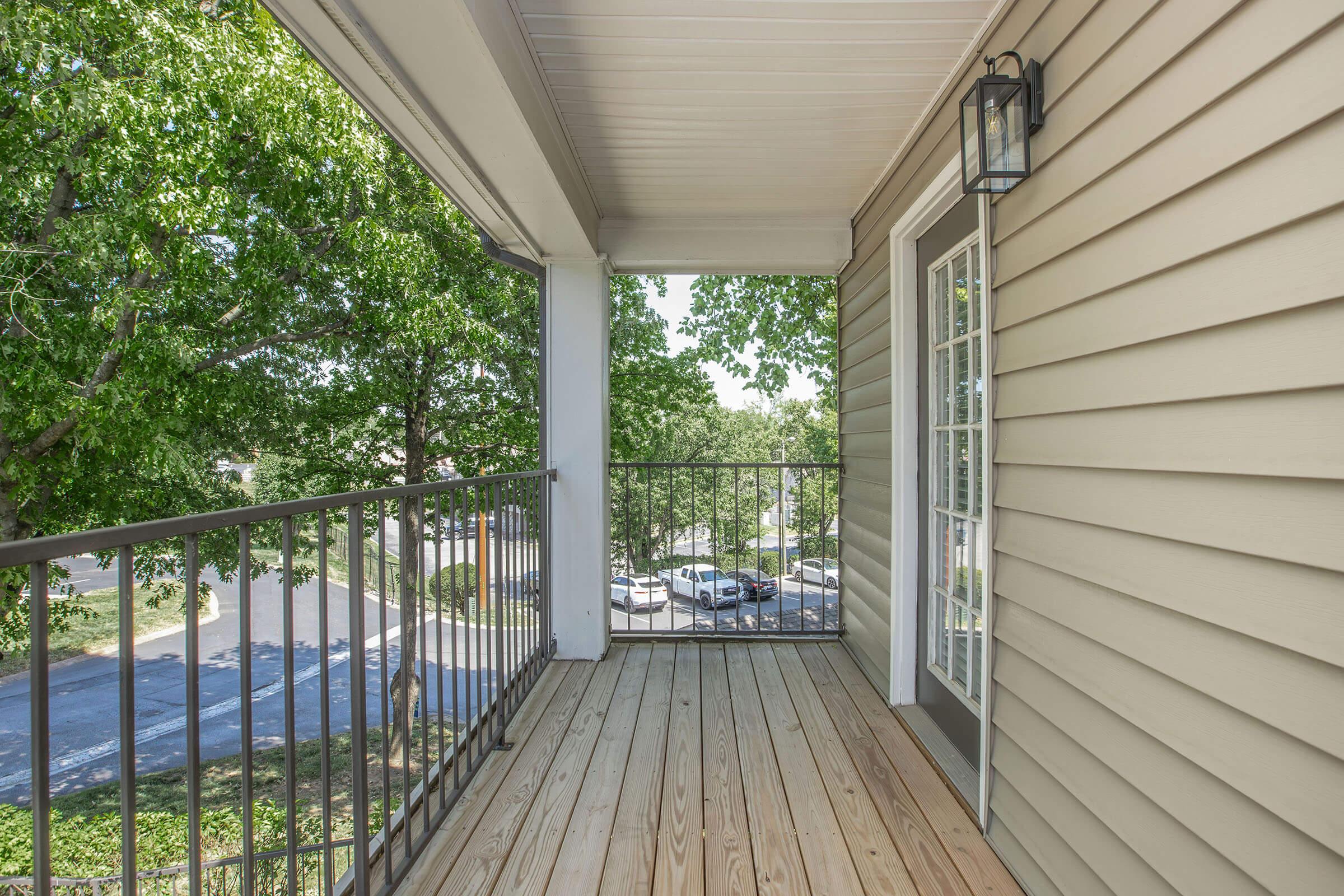 A view from a balcony featuring wooden flooring and a metal railing. The exterior wall is light beige siding, and there's a wall-mounted light fixture. In the background, trees and cars parked on the street are visible, creating a serene outdoor atmosphere.