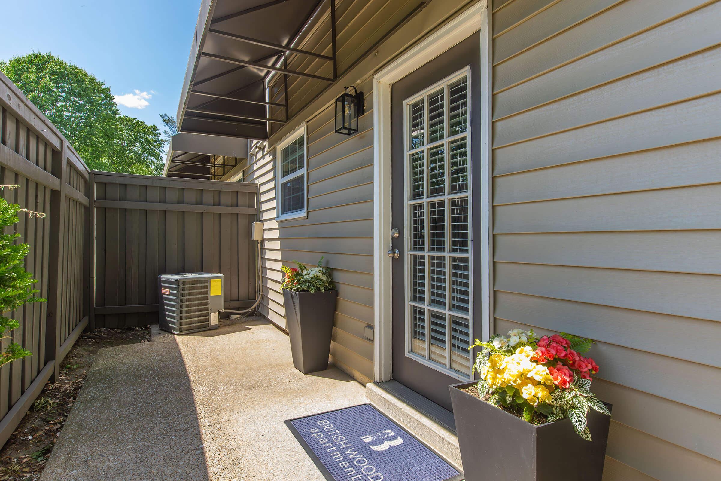A welcoming entrance to a home featuring a gray door with glass panels, potted flowers in front, and a welcome mat. An air conditioning unit is positioned beside the house, surrounded by a fence and greenery. The scene is bright and inviting, indicating a well-maintained outdoor space.