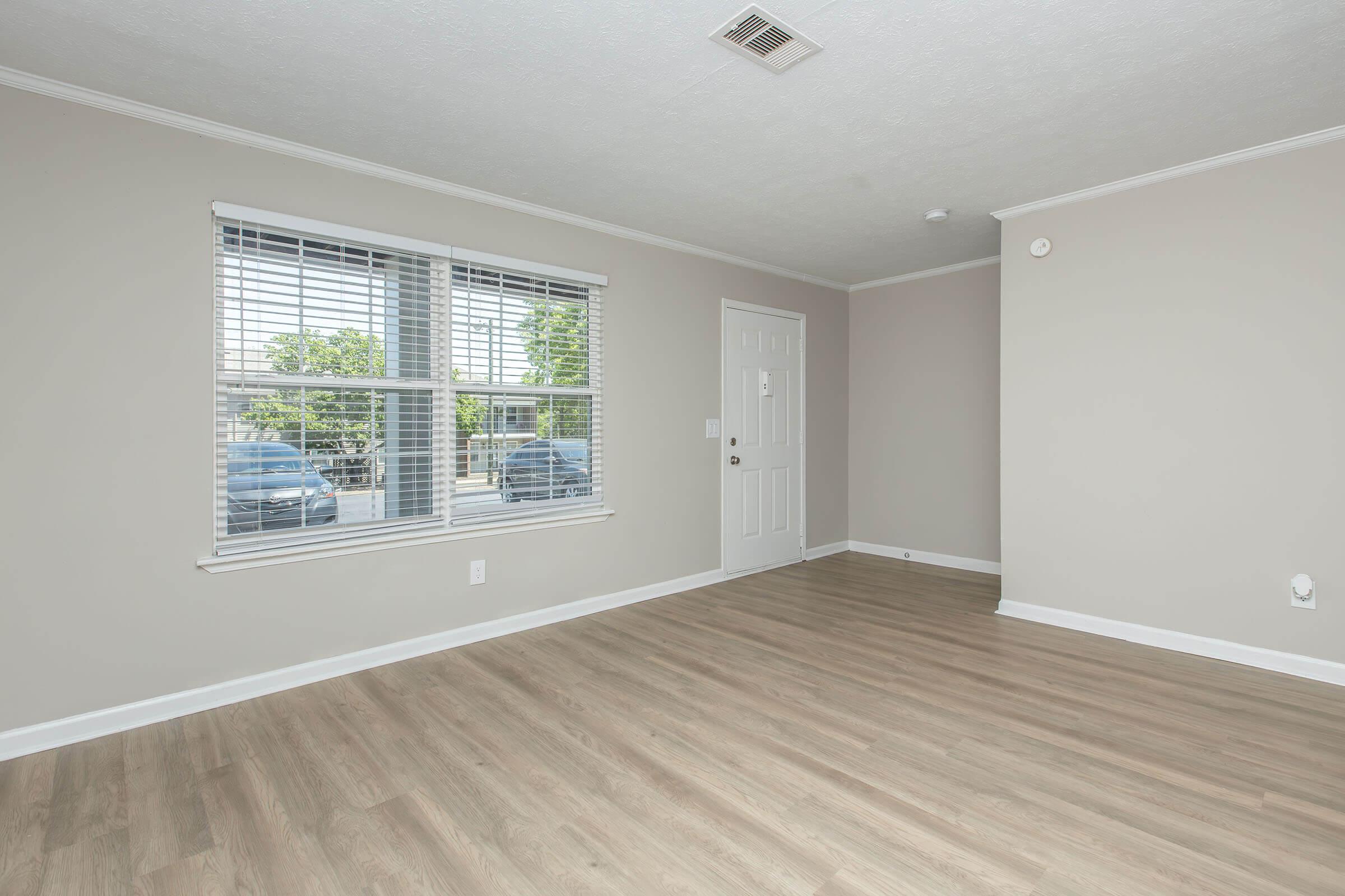 A vacant room with light-colored walls and laminate flooring. Two large windows allow natural light, and a white door is visible on one wall. The space is clean and unfurnished, creating a bright and open atmosphere.
