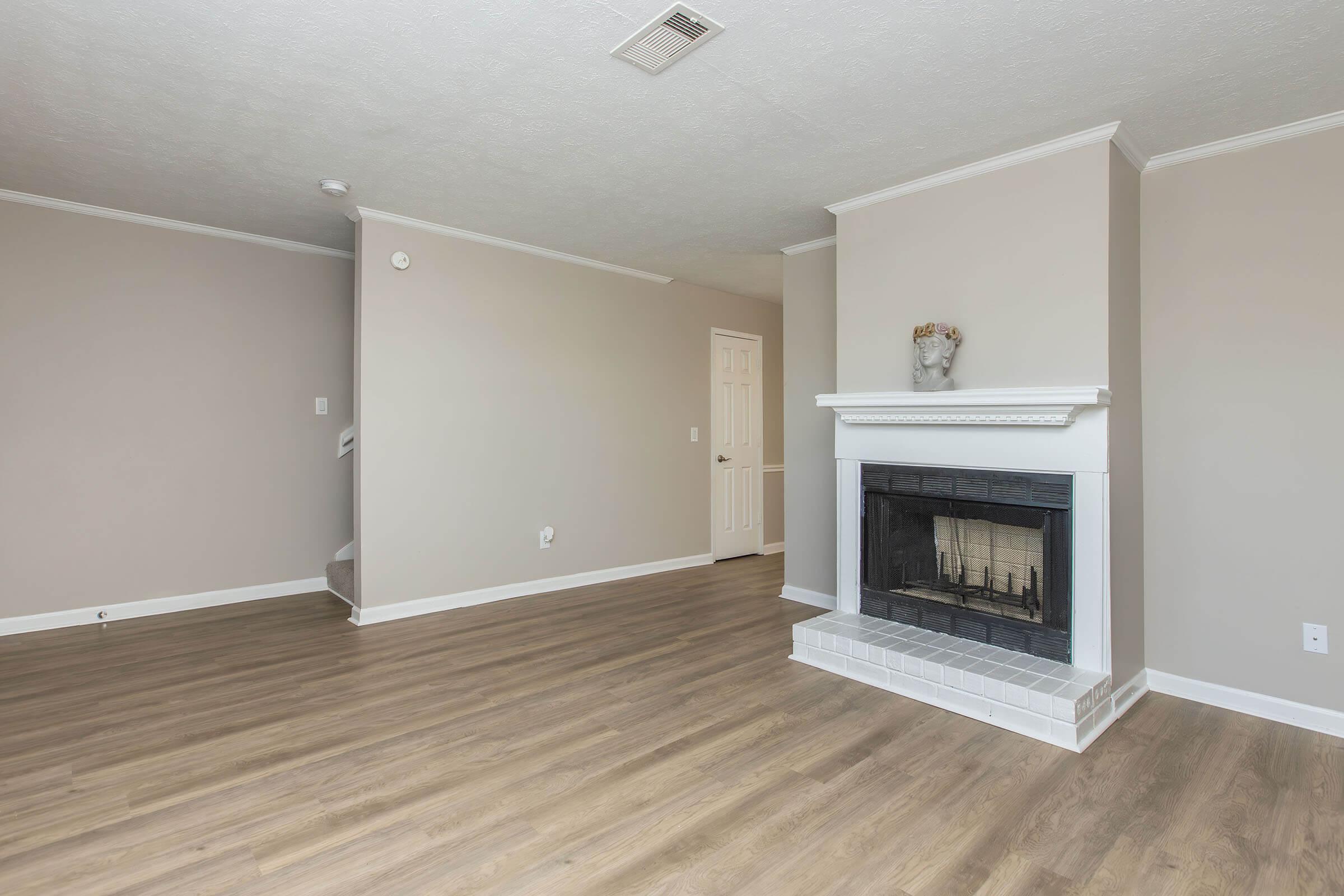 An empty living room featuring light-colored walls, hardwood flooring, and a black fireplace with a white mantel. There is a small decorative item on the mantel, and a doorway leads to another room. A staircase is visible on the left side, and the space is well-lit with natural light.
