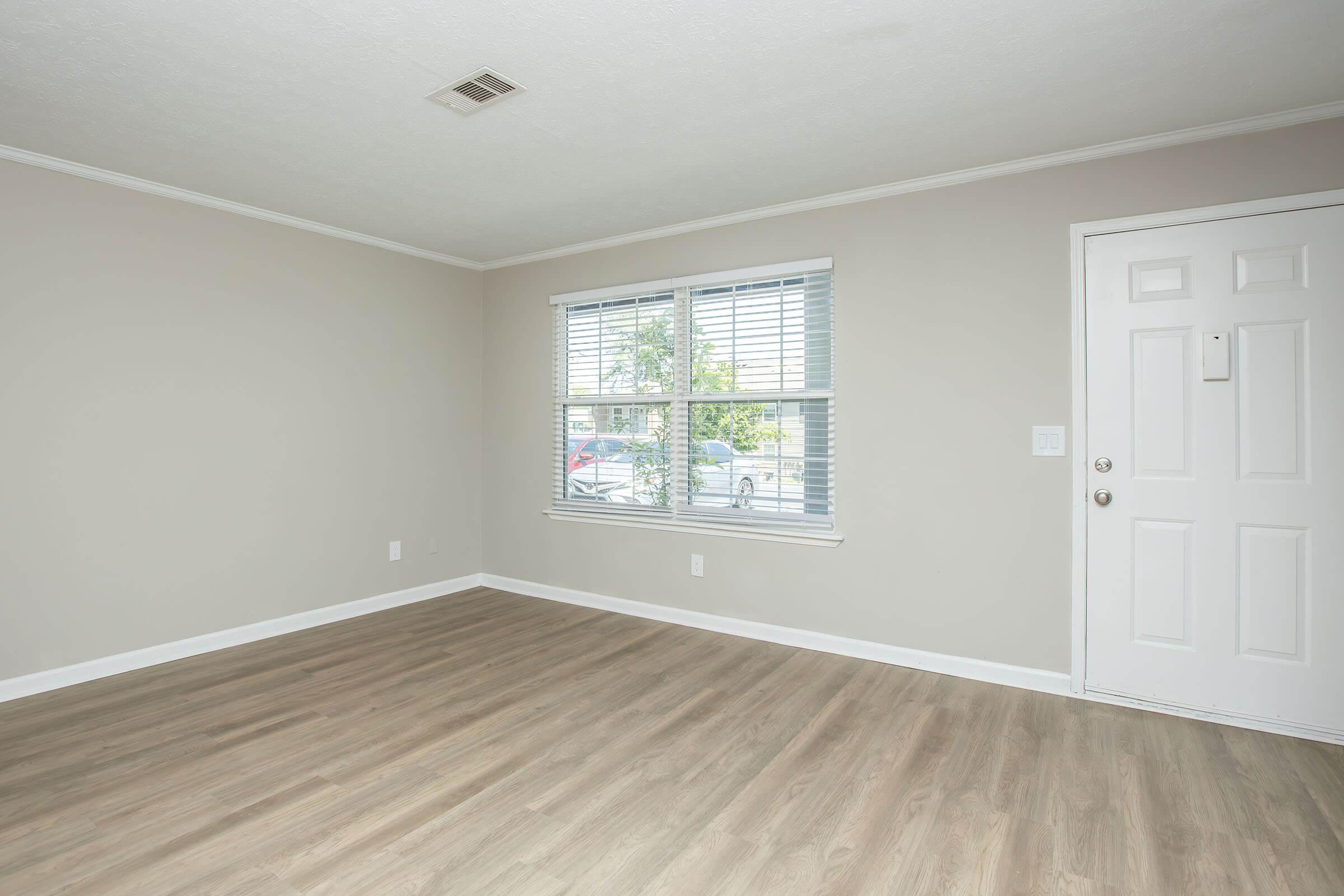 A bright and empty room with light gray walls and wooden flooring. It features a large window with white blinds, letting in natural light, and a door to the right. The space is minimalistic, with no furniture or decorations, providing a clean and open atmosphere suitable for various uses.