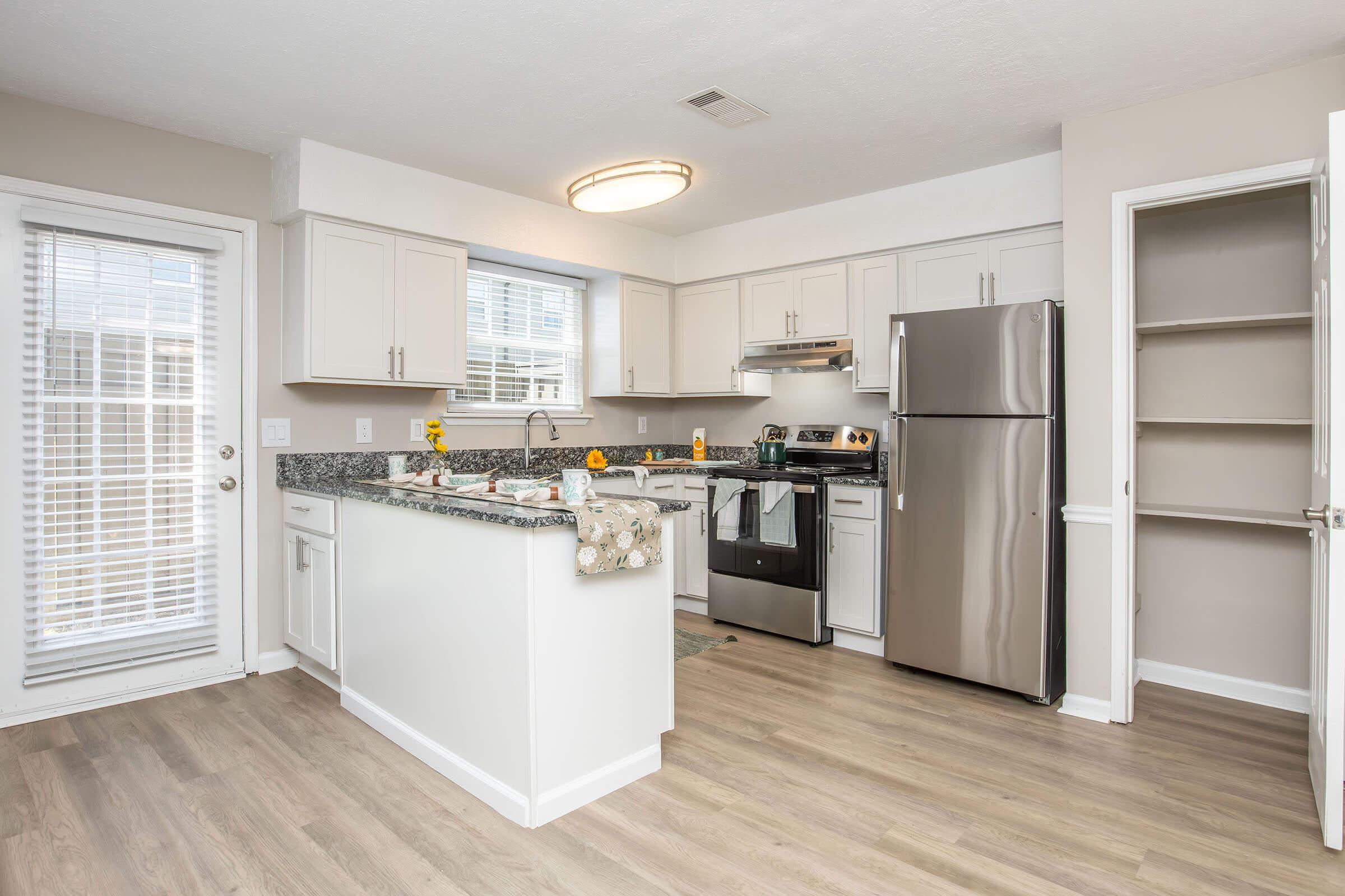 A modern kitchen featuring light-colored cabinetry, granite countertops, and stainless steel appliances. There is a window above the sink with a view to the outside and a sliding glass door leading to a patio. The flooring is light wood, and there is a pantry to the right.