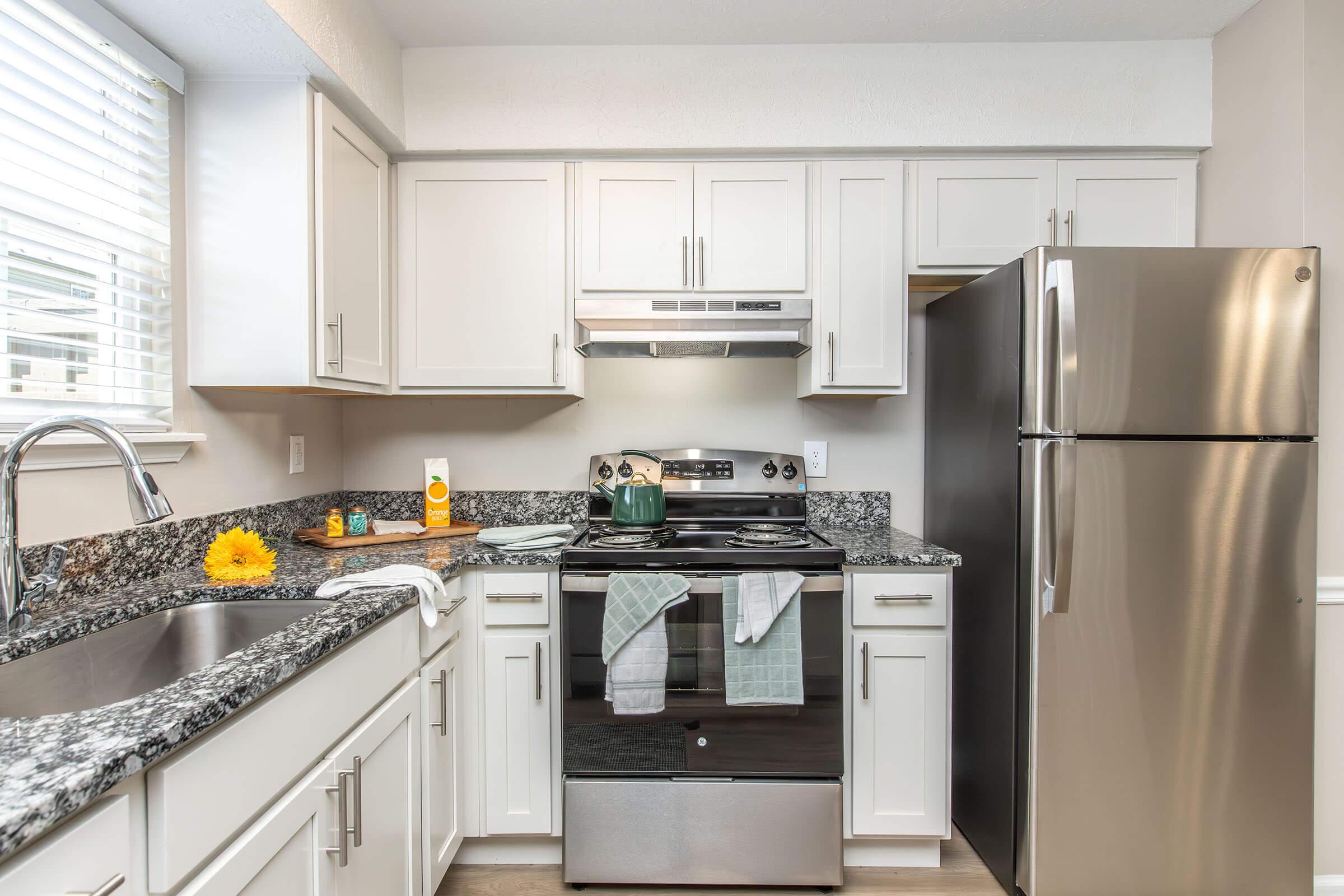 Modern kitchen featuring white cabinetry, granite countertops, a stainless steel refrigerator, and a black stove with a built-in microwave above. Natural light enters through a window, and small kitchen accessories, including a sunflower decoration, are visible on the countertop.