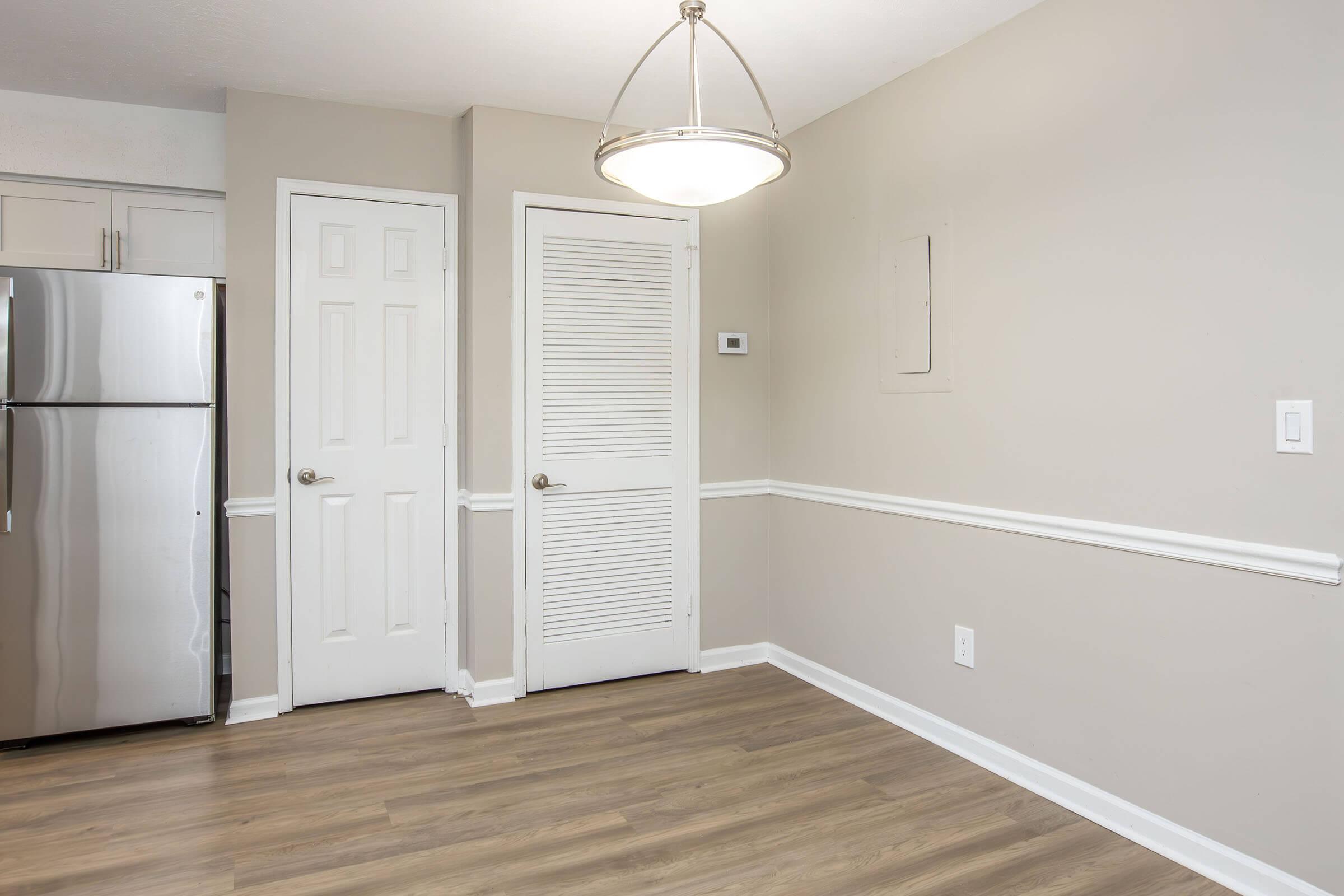 A well-lit kitchen area featuring a stainless steel refrigerator, light wood flooring, and smooth beige walls. Two doors are visible: one leads to a closet and the other is a standard door. A ceiling light fixture hangs above, providing additional illumination. The space appears modern and tidy.