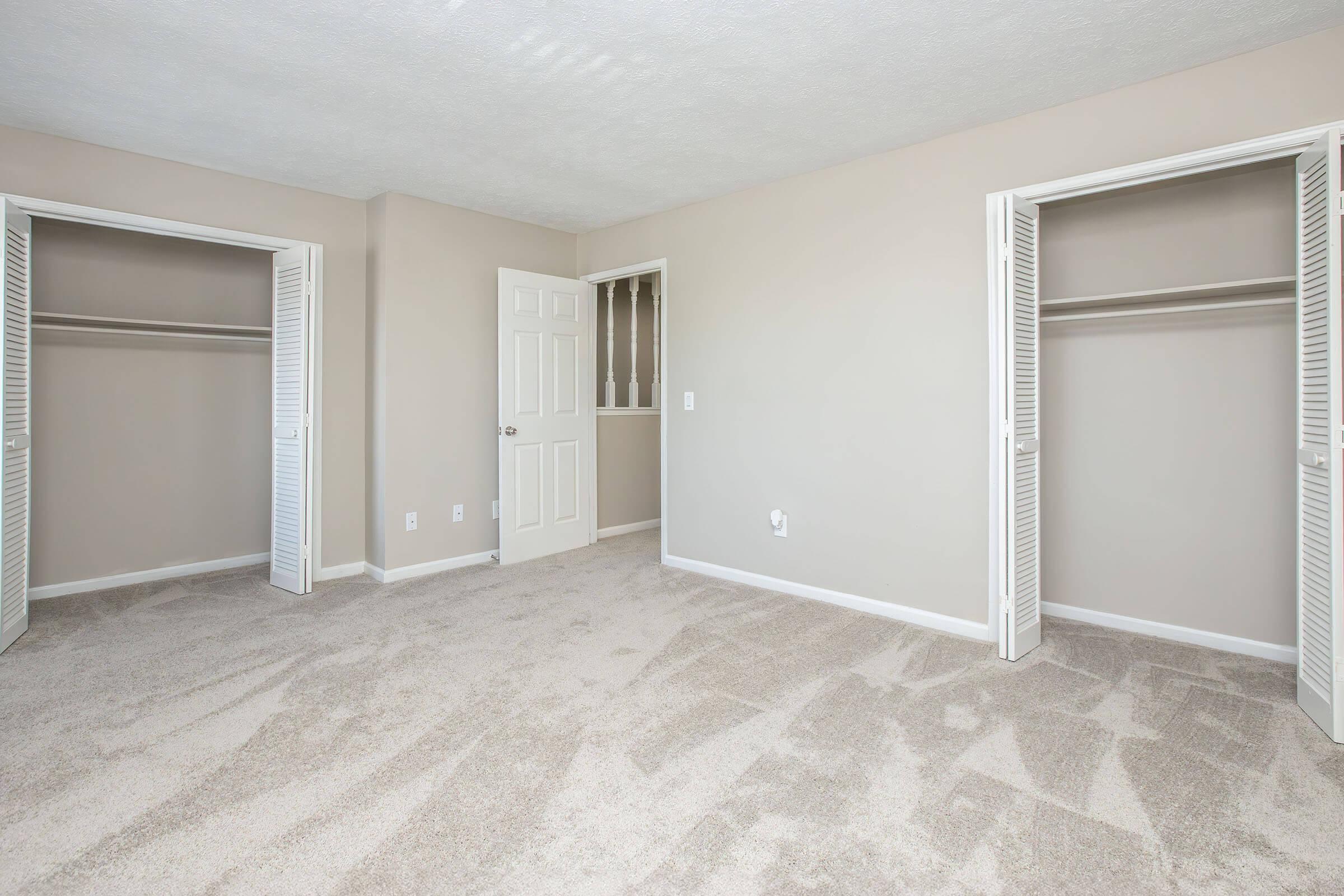 A spacious, empty bedroom featuring light beige walls, plush carpet flooring, and two closet doors. A single door leads to another room, with ample natural light enhancing the open space. The overall ambiance is clean and inviting, perfect for customization.
