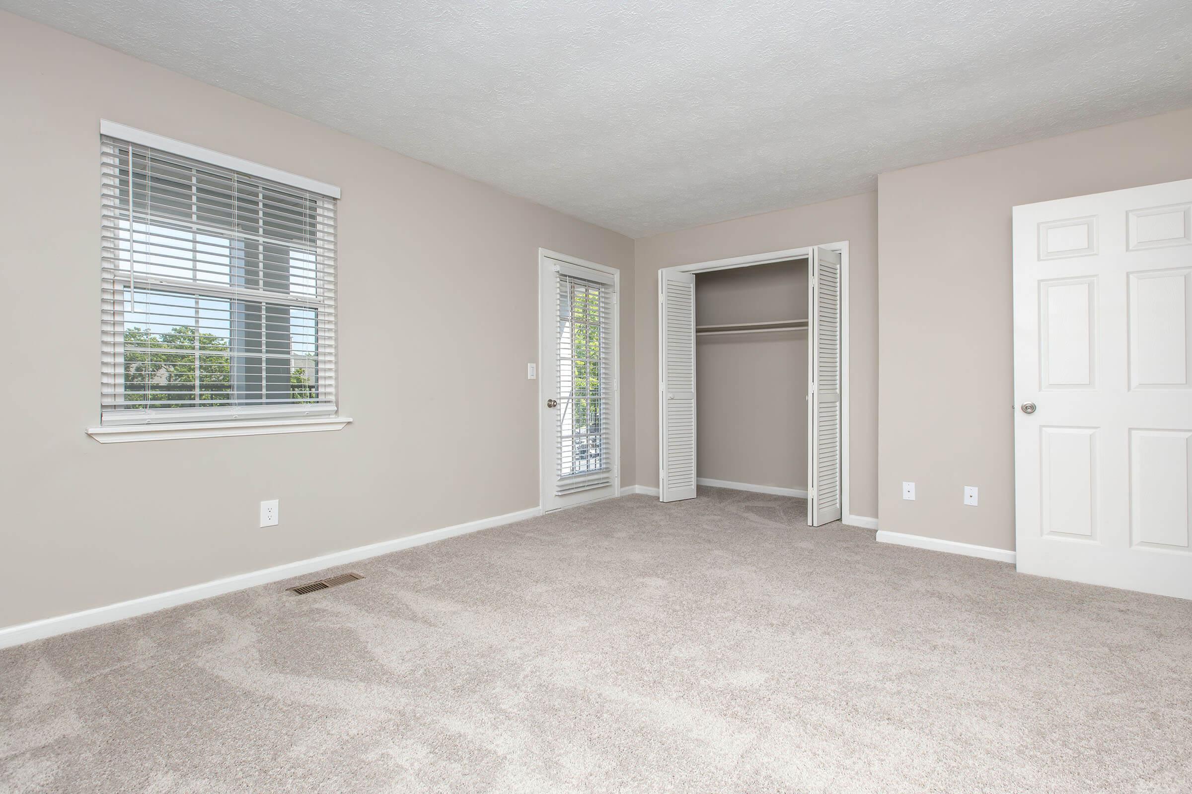 A well-lit, empty room featuring light beige walls and carpet. There is a window with blinds on the left, a sliding door leading to a balcony, and a closet with double doors. The overall ambiance is calm and spacious, ideal for personal decoration or furnishing.
