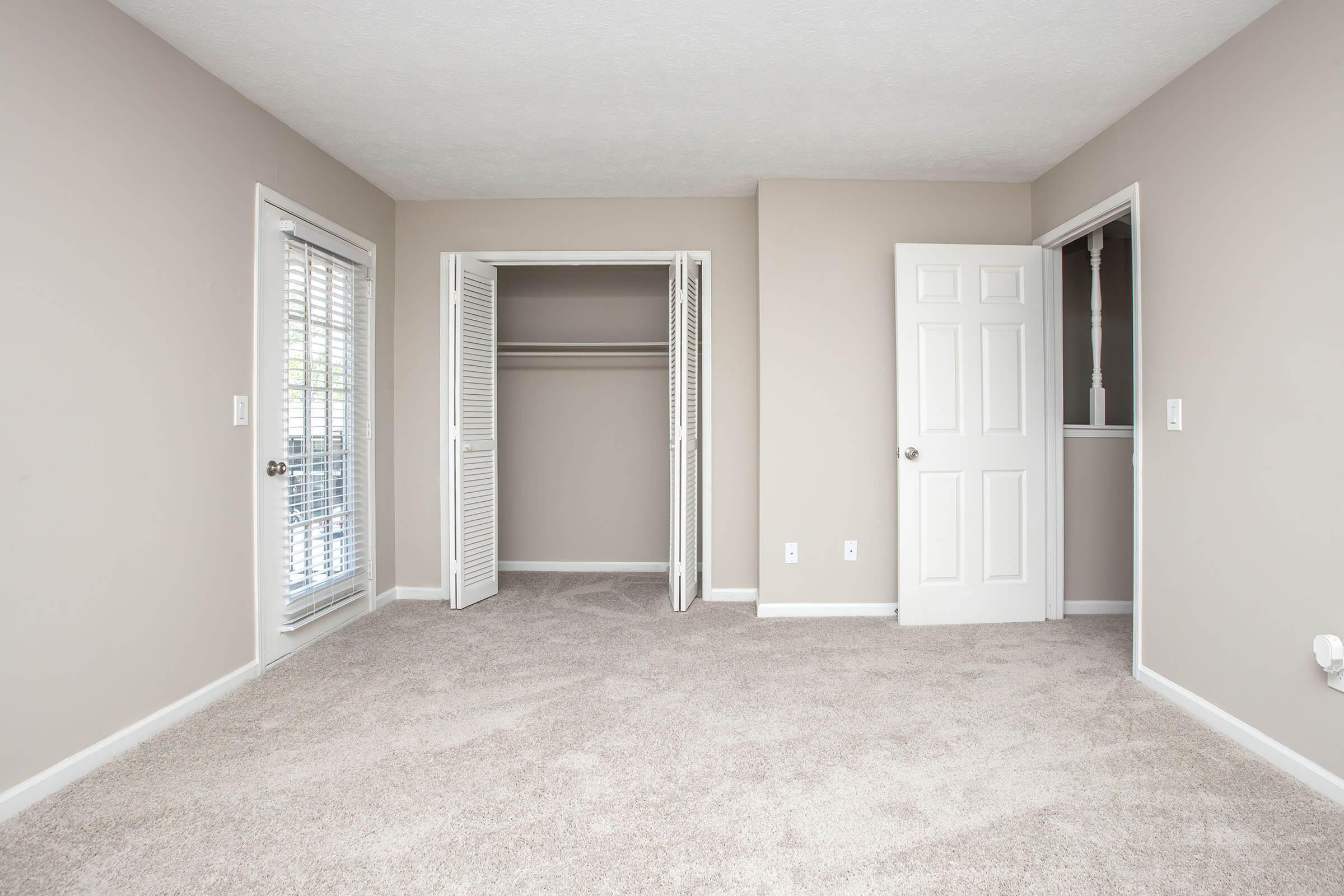 A spacious, light-colored bedroom featuring beige walls and carpeted flooring. There are two doors: one leads to a closet with sliding doors, and the other opens to a small room or hallway. Natural light enters through a window with blinds. The overall atmosphere is clean and inviting.