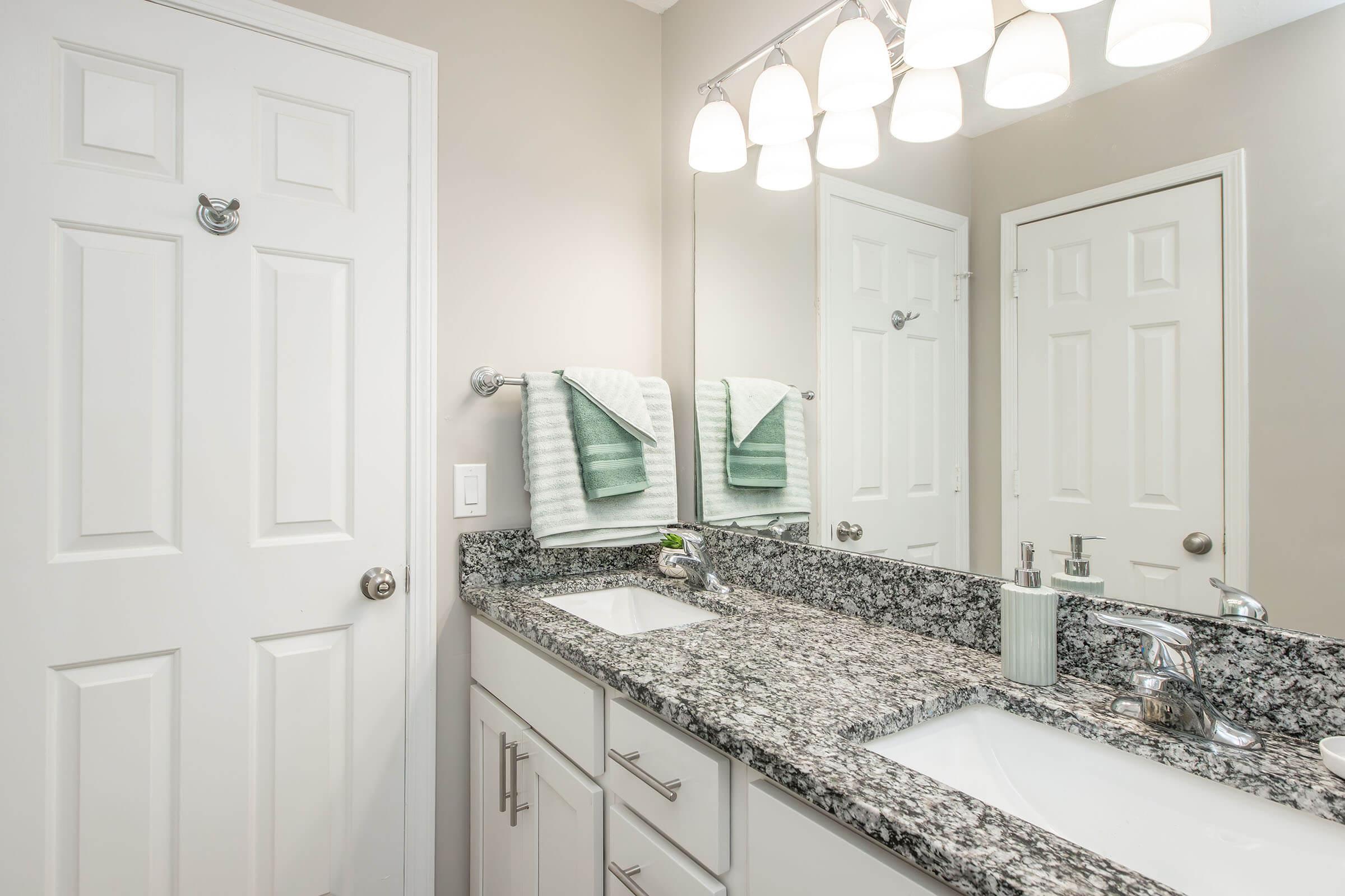 A modern bathroom featuring a double vanity with a granite countertop, two sinks, and stylish faucets. There are white cabinets and a mirror with bright lighting above. Two green towels hang neatly on towel bars, and a white door is partially visible in the background.
