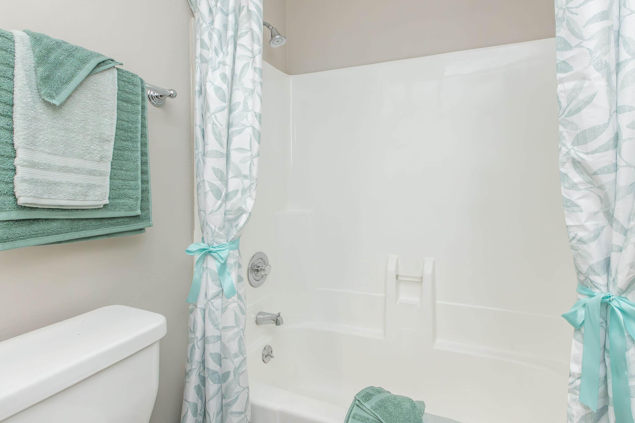 A clean, modern bathroom featuring a bathtub with a white shower curtain adorned with a leaf pattern. There are teal towels hanging on a rack and neatly folded towels on the tub's edge, adding a pop of color to the neutral walls and fixtures. Decorative accents enhance the overall fresh and inviting atmosphere.