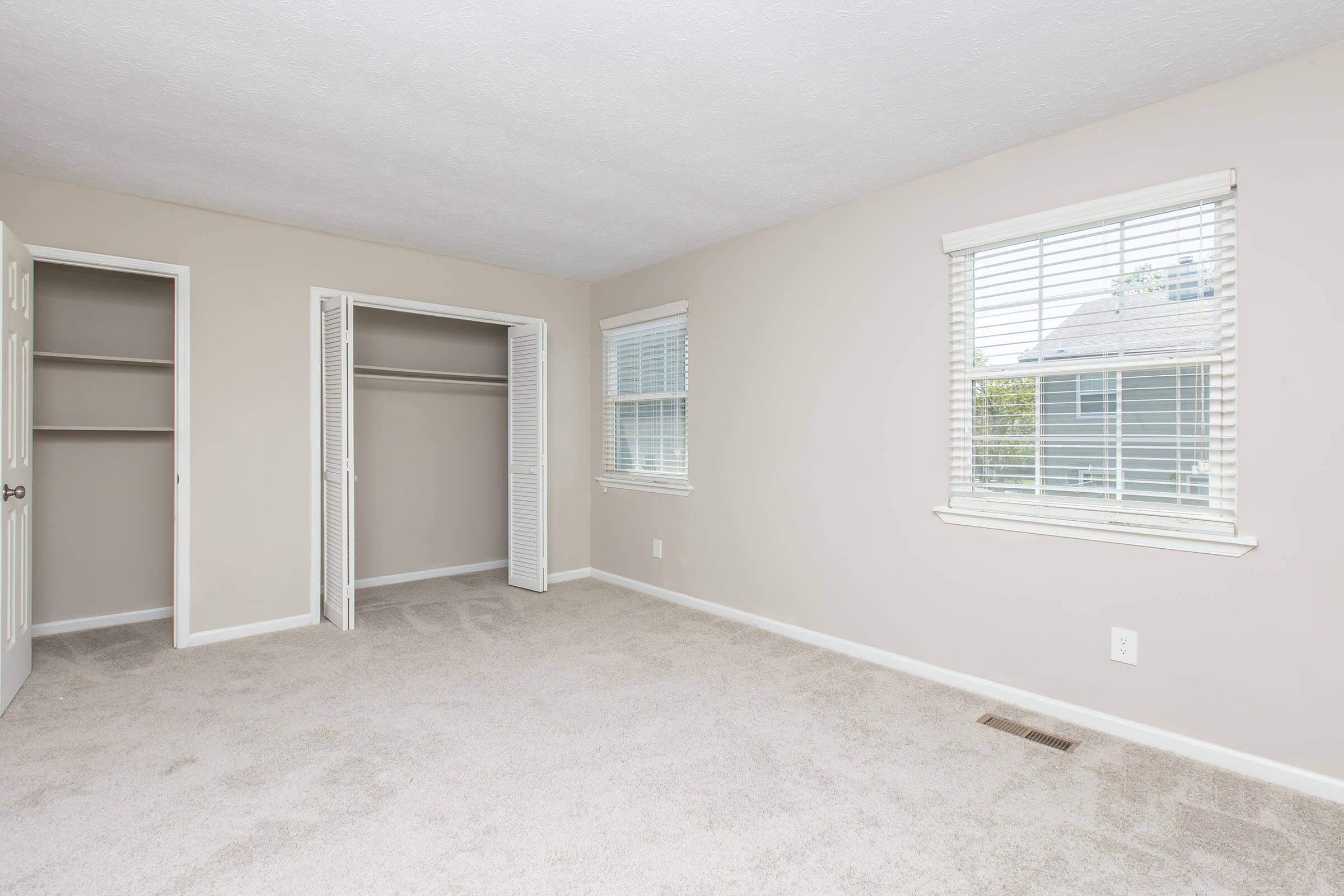A spacious, empty room featuring light beige walls and carpet. It has a large closet with double doors on one side and two windows that let in natural light. The room appears bright and airy, with a minimalist design.
