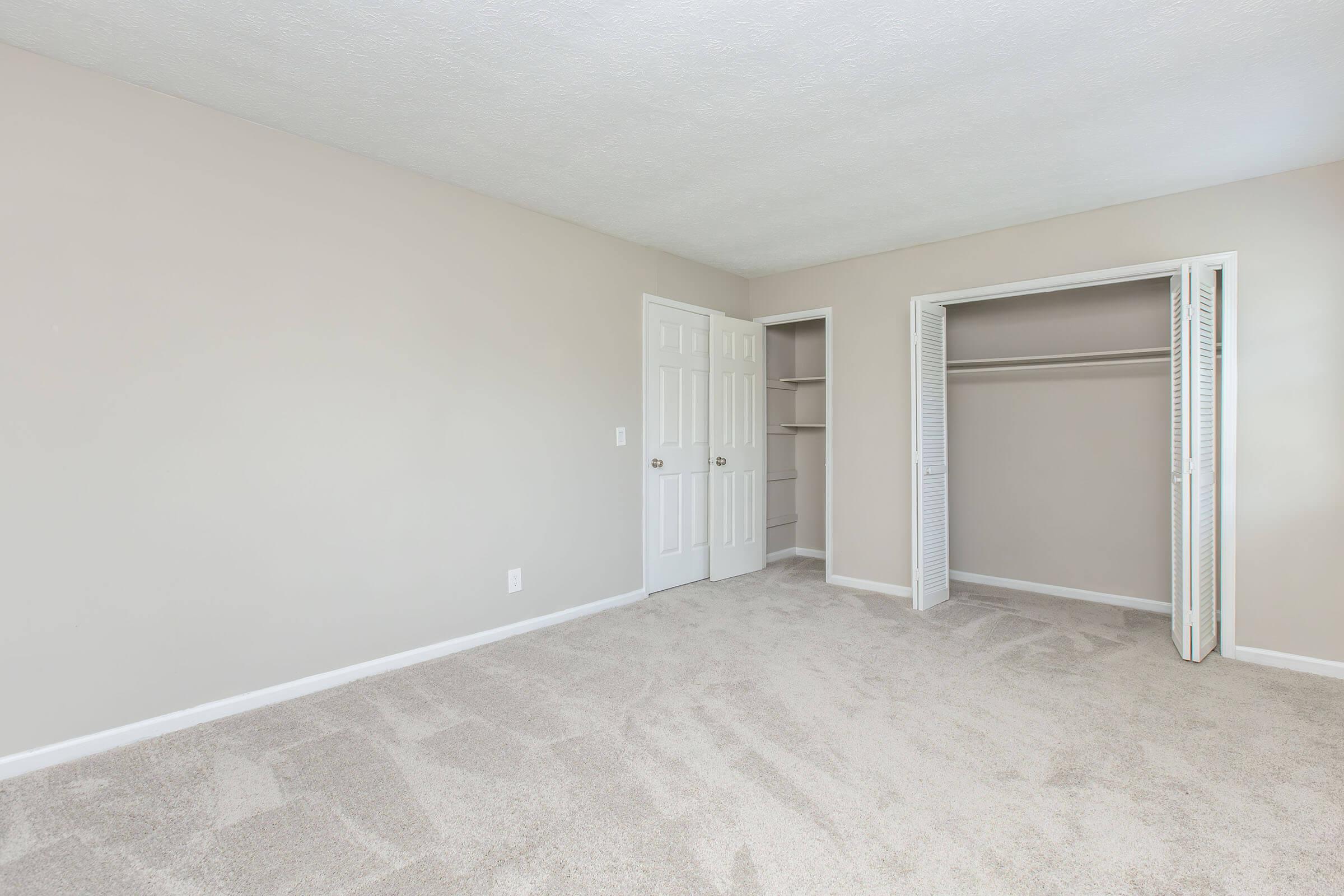 Empty bedroom with light beige walls and gray carpet. There are two closed white doors, one leading to a small closet with sliding doors. The room is well-lit, showcasing its spaciousness and clean, minimalistic design.