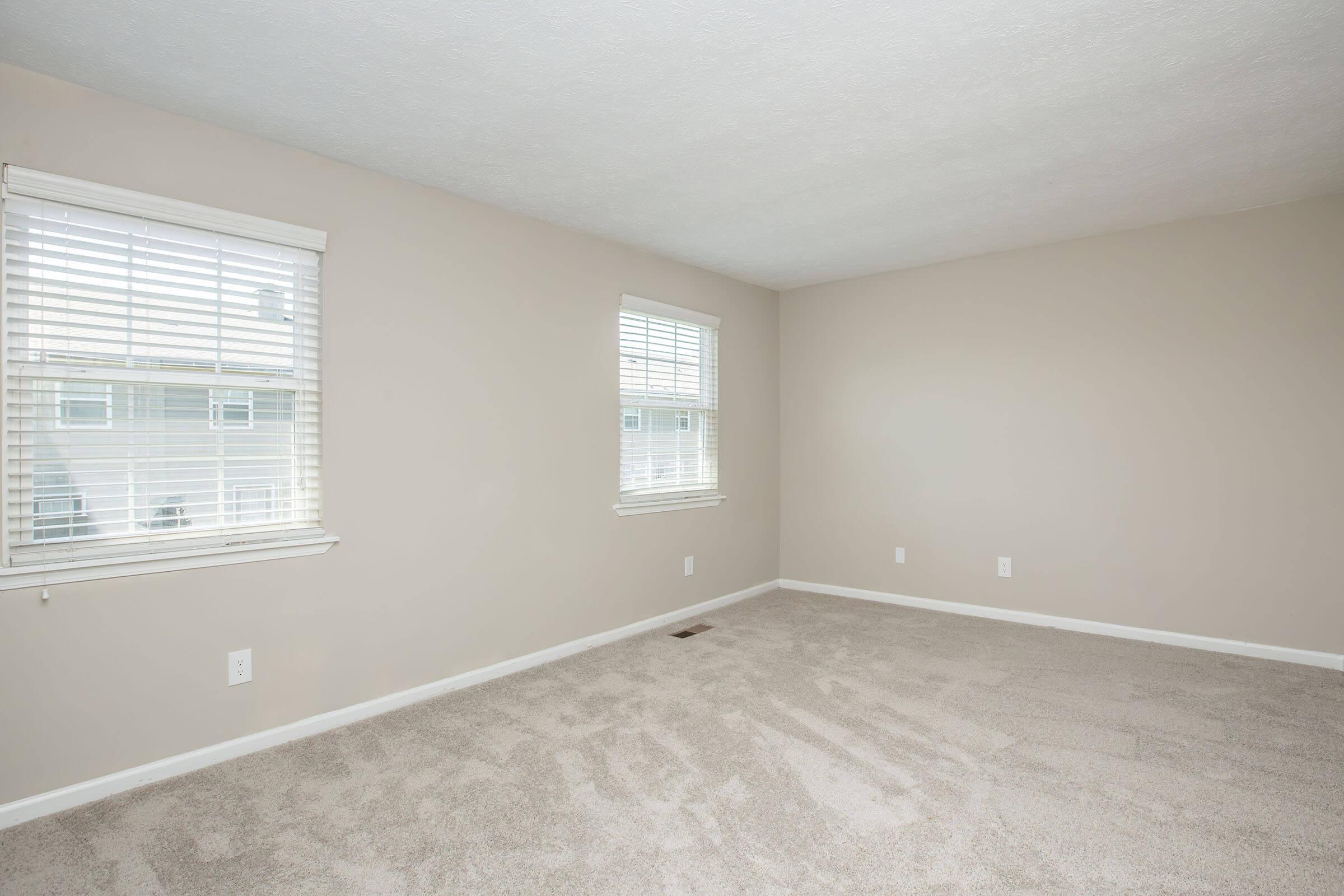 Empty room with light beige walls and plush carpet. Two windows with white blinds allow natural light in, creating a bright atmosphere. Floor is clear, suggesting the space is ready for furniture or decorations. The room has a clean, modern feel, ideal for various uses.