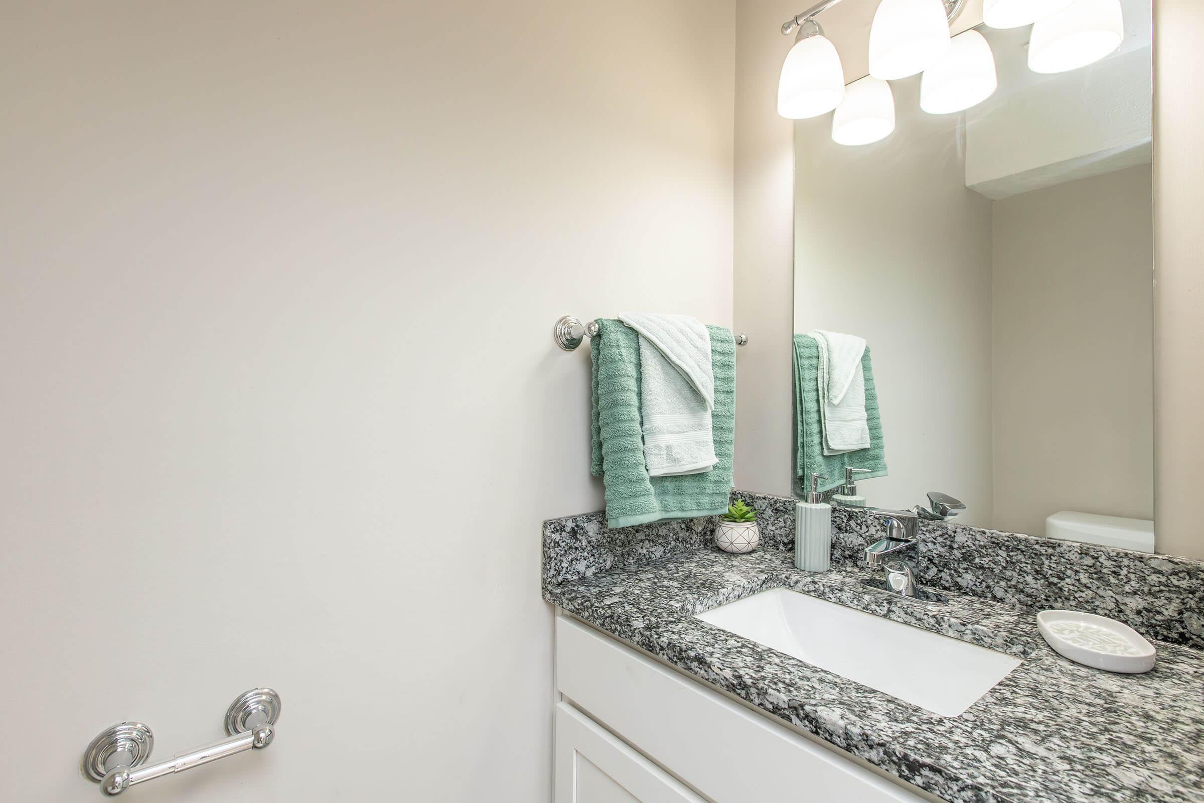 A modern bathroom with a granite countertop featuring a white sink. On the countertop are two light green towels, a small potted plant, and a decorative dish. Above the sink, there is a large mirror and three pendant lights illuminating the space. A towel bar is visible on the wall.