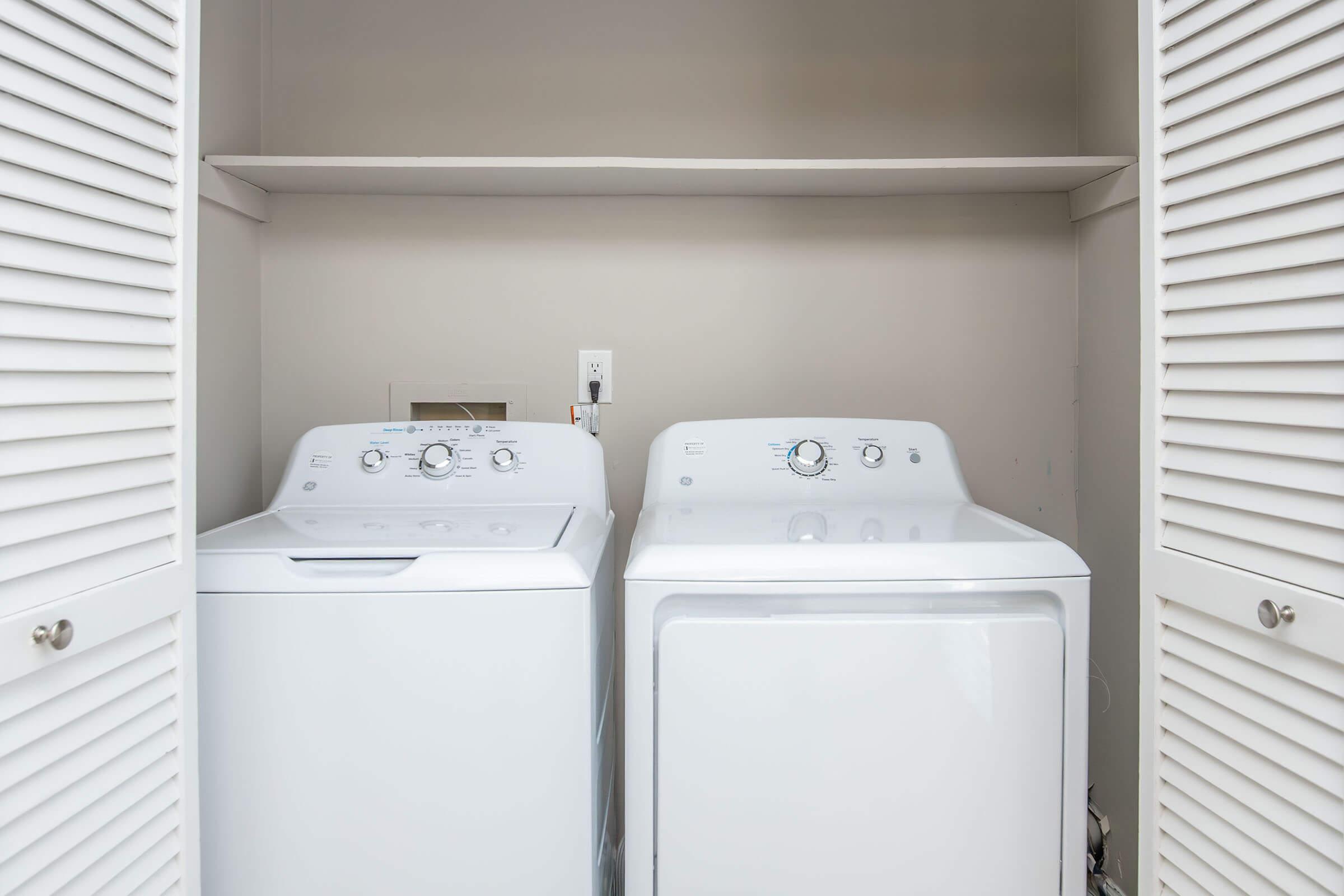 A white washing machine and dryer side by side in a small laundry area, enclosed by white louvered doors. The machines feature control knobs and panels, and a shelf is positioned above them. The background is a light neutral color.