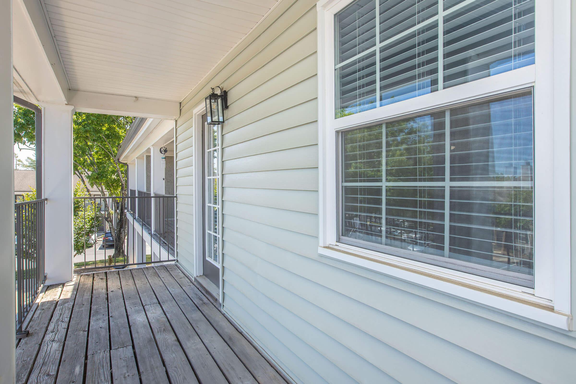 A view of a wooden balcony or porch with a light gray siding exterior. The area features a window with horizontal blinds and a metal railing alongside. The scene is bright, suggesting a sunny day, with greenery visible in the background.