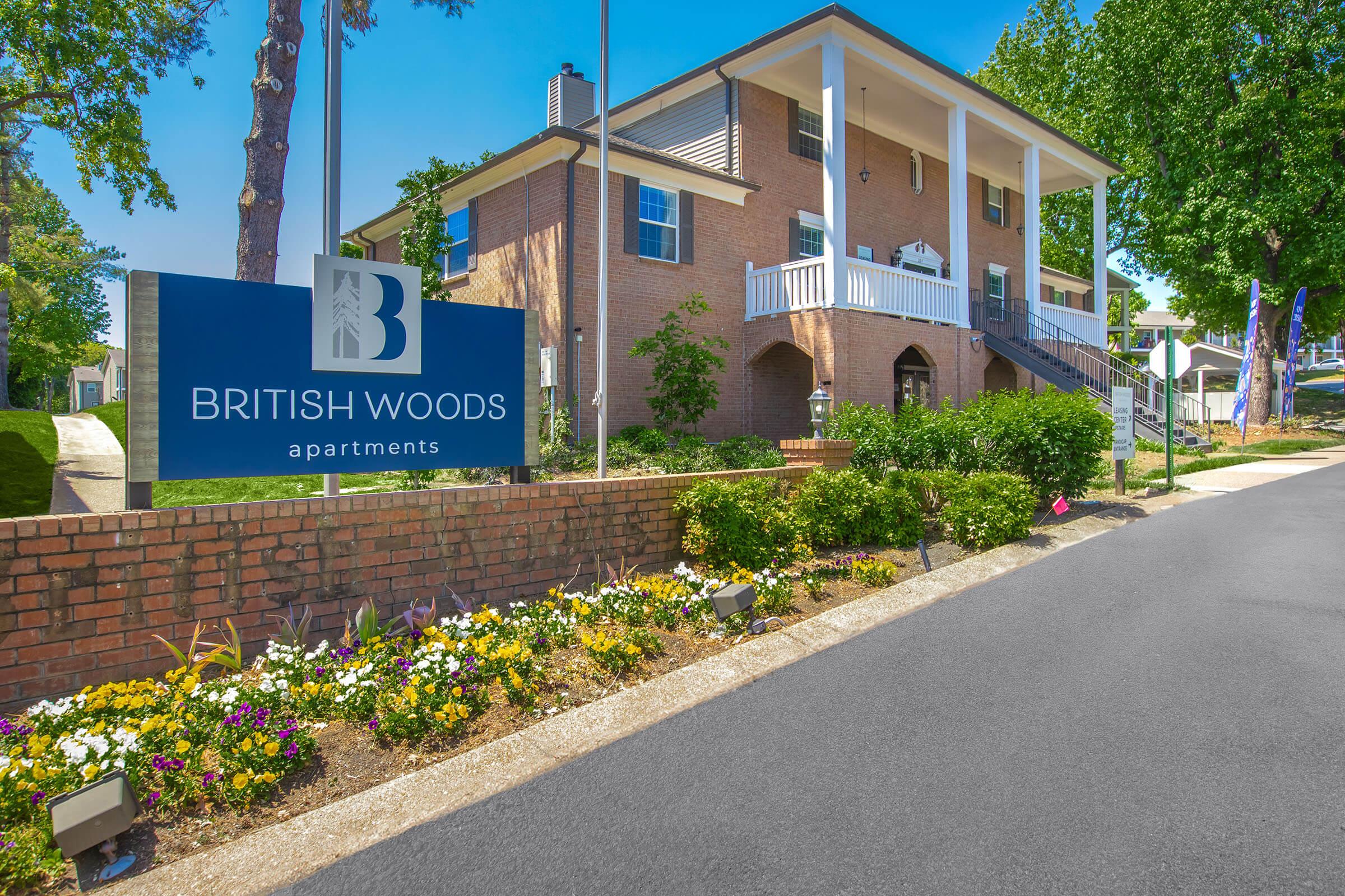 View of the British Woods Apartments, featuring a brick building with a welcoming entrance and a sign in front. The surrounding landscape includes colorful flower beds, a clear blue sky, and trees, creating a bright and inviting atmosphere. Pathway leads towards the entrance of the apartments.