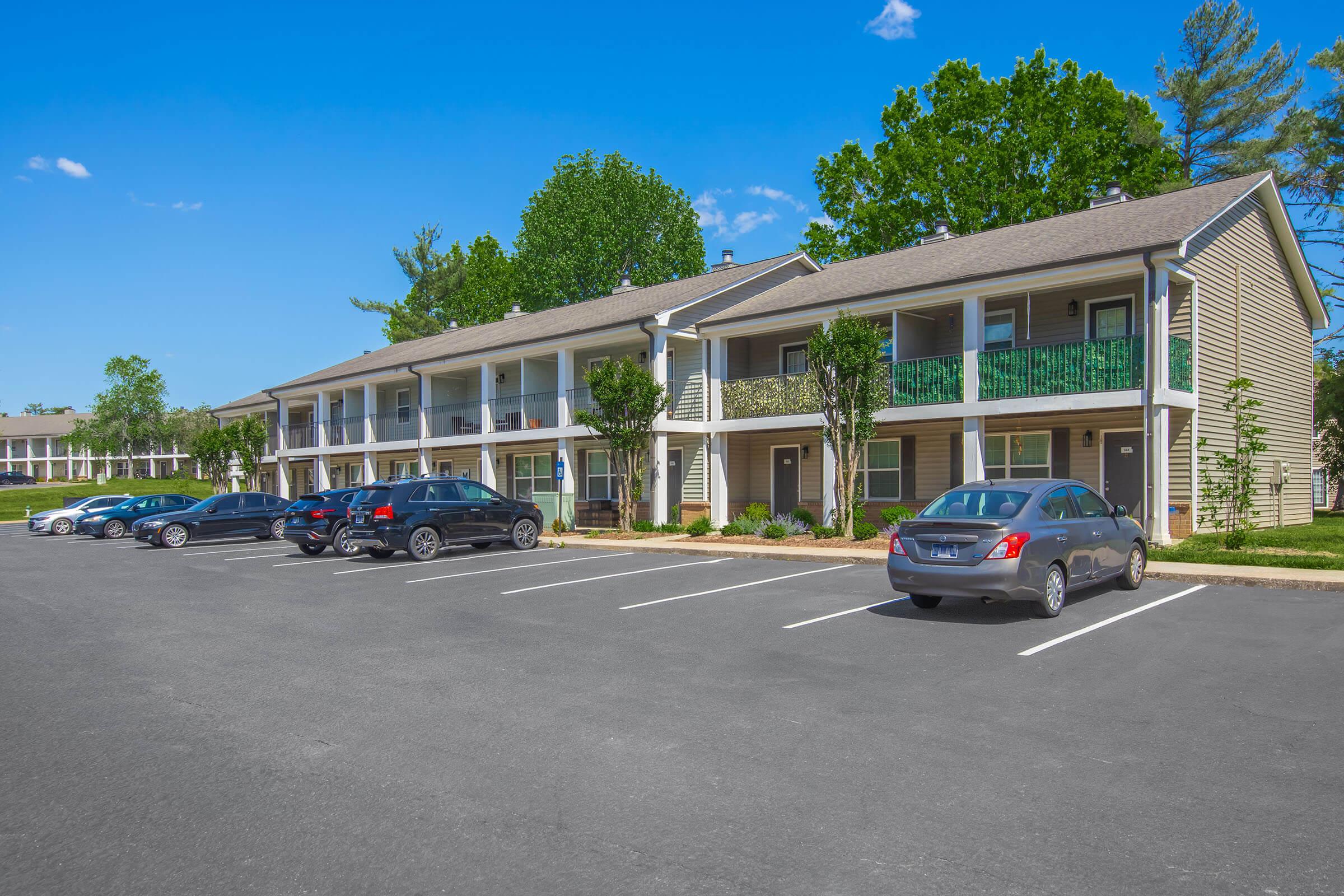 A view of a two-story apartment complex with balconies, surrounded by greenery. Multiple cars are parked in the foreground, with clear blue skies and trees in the background, suggesting a sunny day.