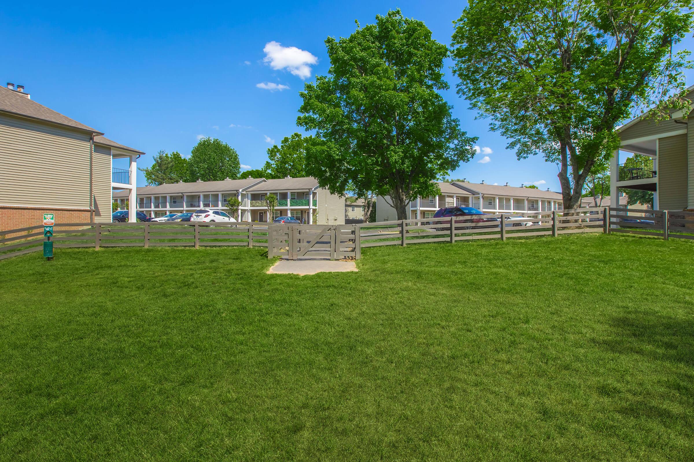 A well-maintained grassy area surrounded by wooden fencing, with trees and blue skies. In the background, there are two apartment buildings and parked cars visible. The scene suggests a residential community with outdoor space for residents.