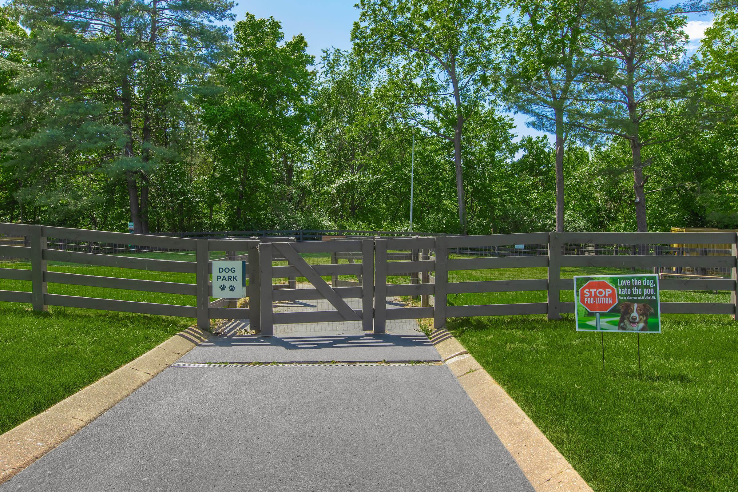 A pathway leading to a fenced dog park entrance, with signs indicating "Dog Park" and "Stop." Lush green grass surrounds the area, and trees are visible in the background, indicating a natural setting suitable for pets. The park appears well-maintained and inviting for dogs and their owners.