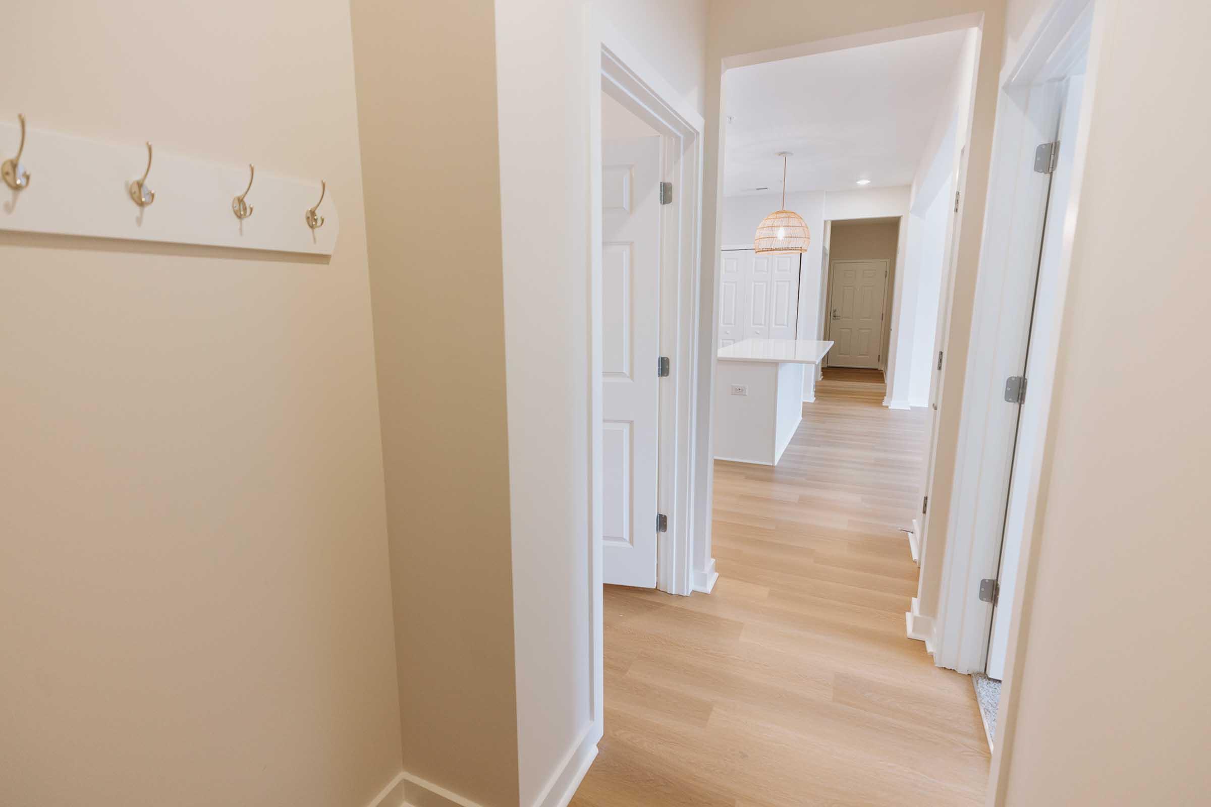 A well-lit hallway featuring light-colored walls and wooden flooring. Coat hooks are mounted on the left wall. Two doors are visible on the right side leading to different rooms, and an open space at the end suggests a bright kitchen area. Soft lighting adds warmth to the entryway.