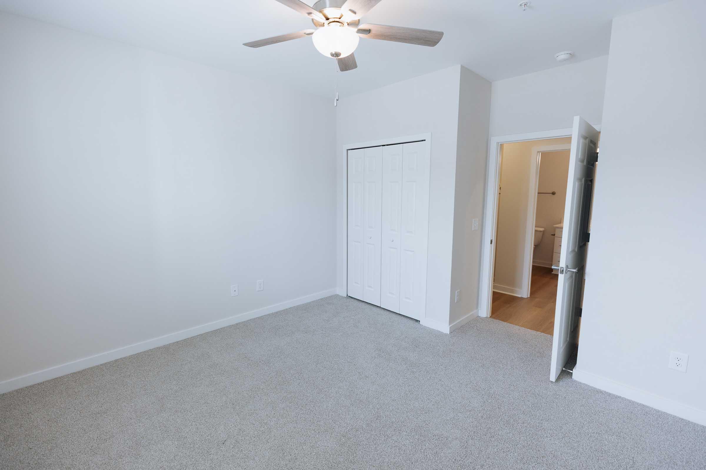 Spacious, empty room with light-colored walls and carpet. A ceiling fan is overhead, and there is a closet with sliding doors on the left. A door on the right leads to a bathroom, visible in the background. The overall atmosphere is bright and airy.