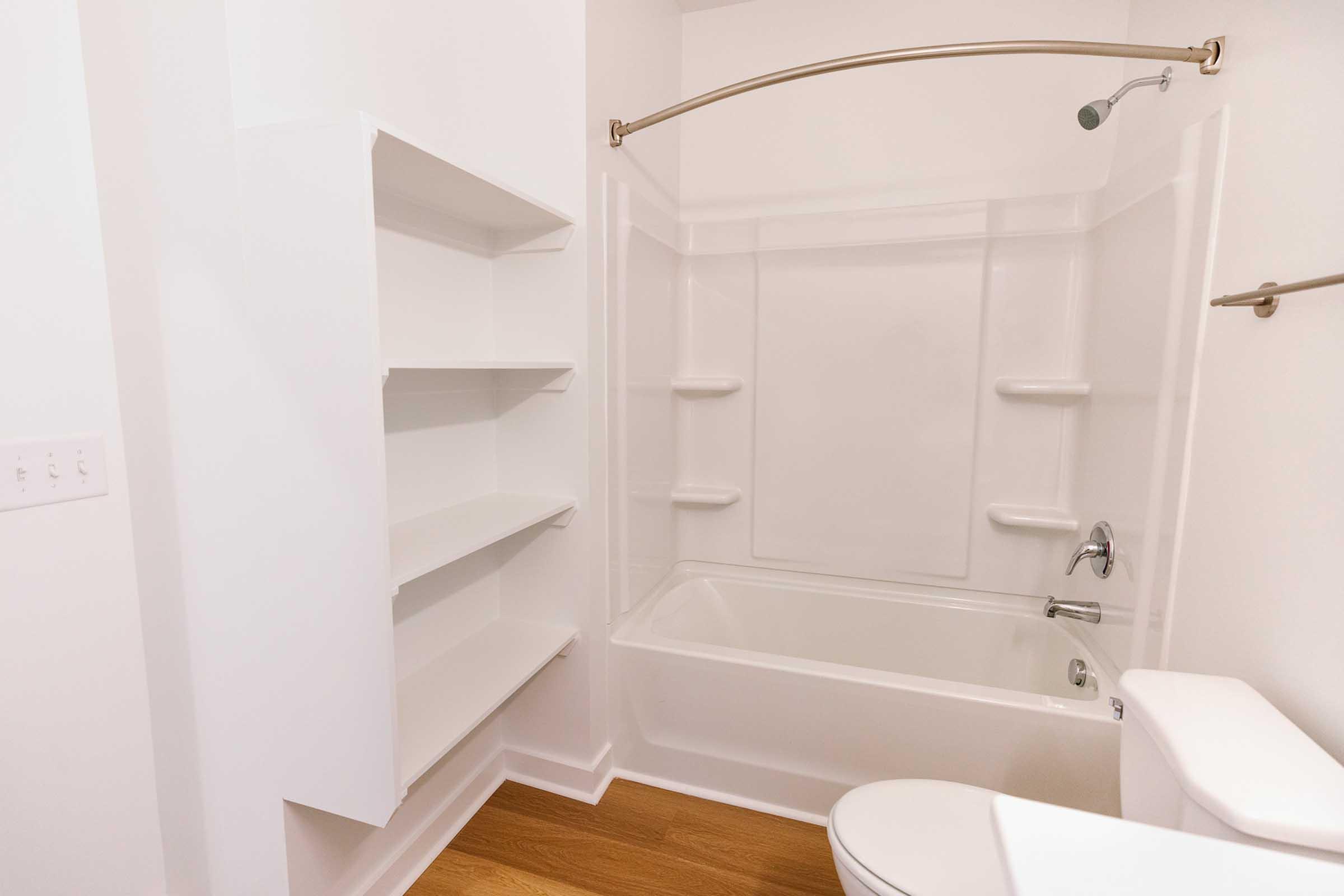 A clean and modern bathroom featuring a bathtub with a showerhead above, white walls, and light wooden flooring. There are built-in shelves on the left side for storage and a white toilet on the right, creating a bright and spacious atmosphere.