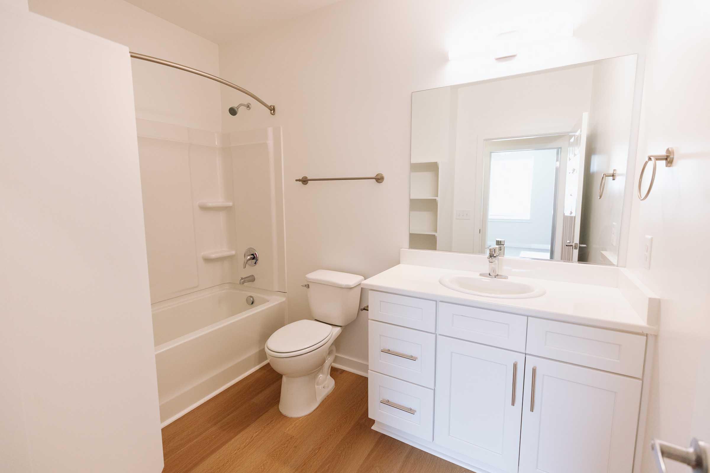 A modern bathroom featuring a white bathtub, toilet, and vanity with a sink. The walls are mostly white, and the floor has light wooden flooring. A large mirror is mounted above the sink, and there is a glass door leading to another area visible in the background. Brightly lit and clean design.