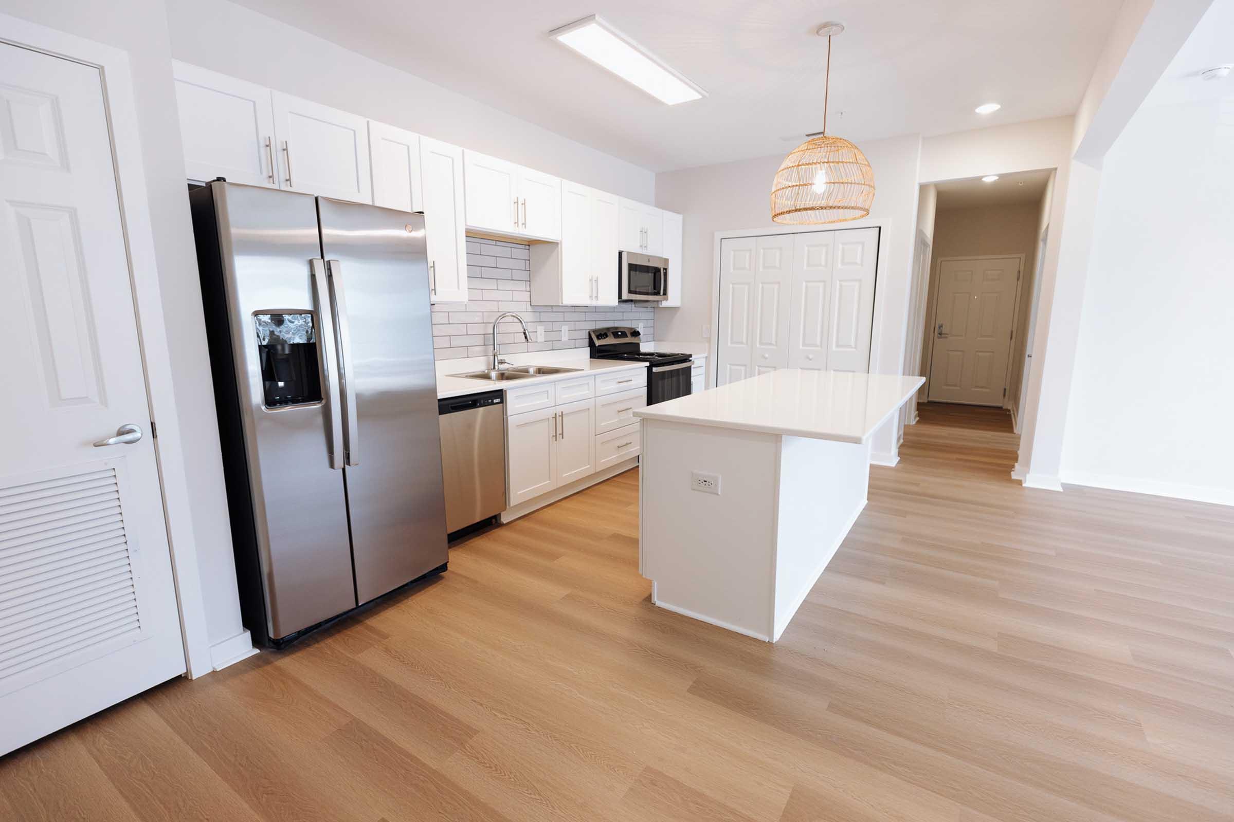 Modern kitchen featuring stainless steel appliances including a refrigerator, oven, and microwave. The space has white cabinetry, a sleek countertop, and a hanging light fixture. The floor is light wood, and the design emphasizes a bright and open concept.