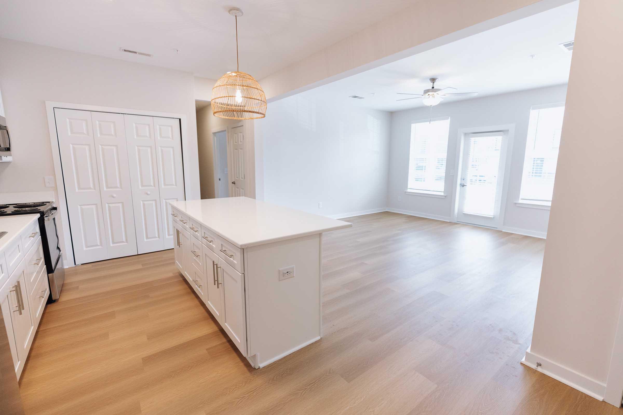 A spacious, modern kitchen and living area with light wood flooring. The kitchen features a white island and cabinetry, with a pendant light hanging above. There are two windows in the living area, allowing natural light to fill the space. The walls are painted in a light color, creating a bright atmosphere.