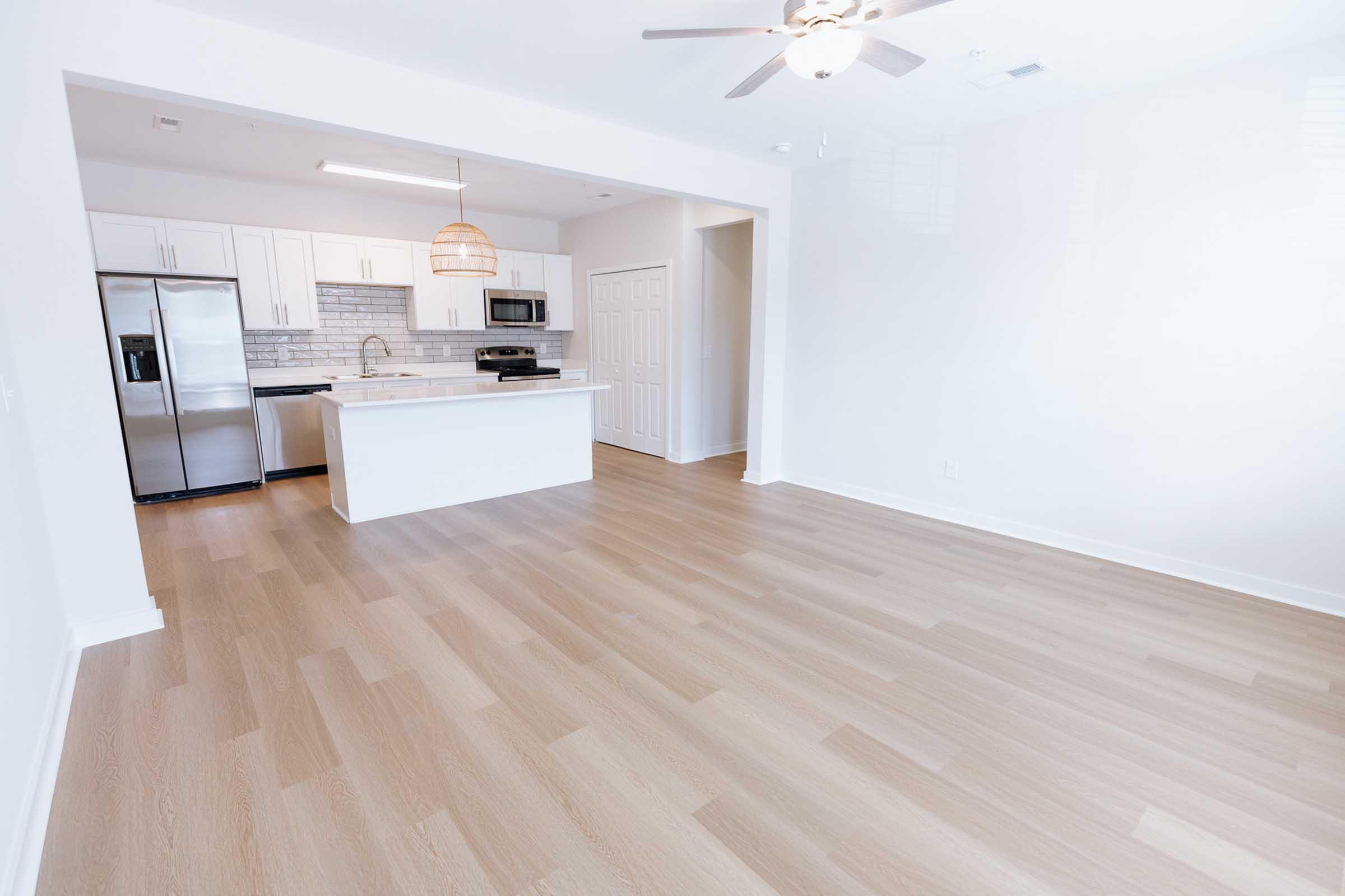 A bright, modern kitchen and living area with light-colored walls and hardwood floors. The kitchen features white cabinets, a black refrigerator, stainless steel appliances, and a light fixture above the island. The open space has a ceiling fan and is well-lit with natural light, creating a welcoming atmosphere.