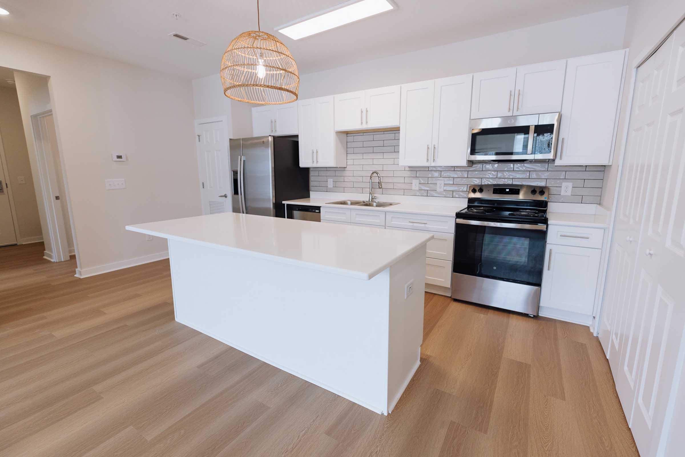 A modern kitchen featuring a large white island, stainless steel appliances, and white cabinetry. Natural light fills the space, illuminating the sleek countertops and backsplash. The flooring is light-colored wood, complementing the fresh, contemporary design.