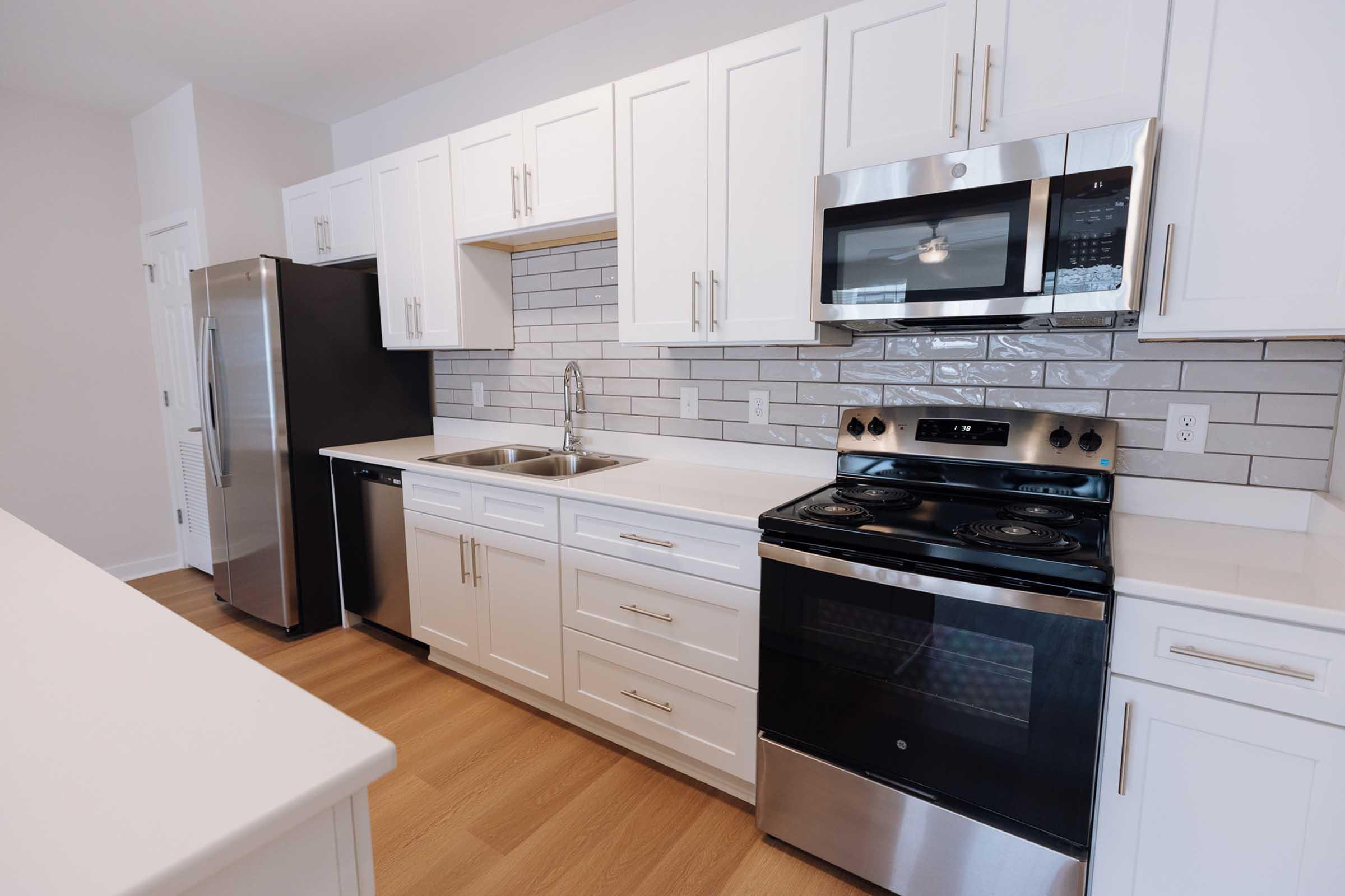 Modern kitchen featuring white cabinetry, stainless steel appliances including a refrigerator, microwave, and oven, a double sink, and a sleek backsplash with gray tiles. The space has light wood flooring and ample counter space, providing a clean and functional cooking area.