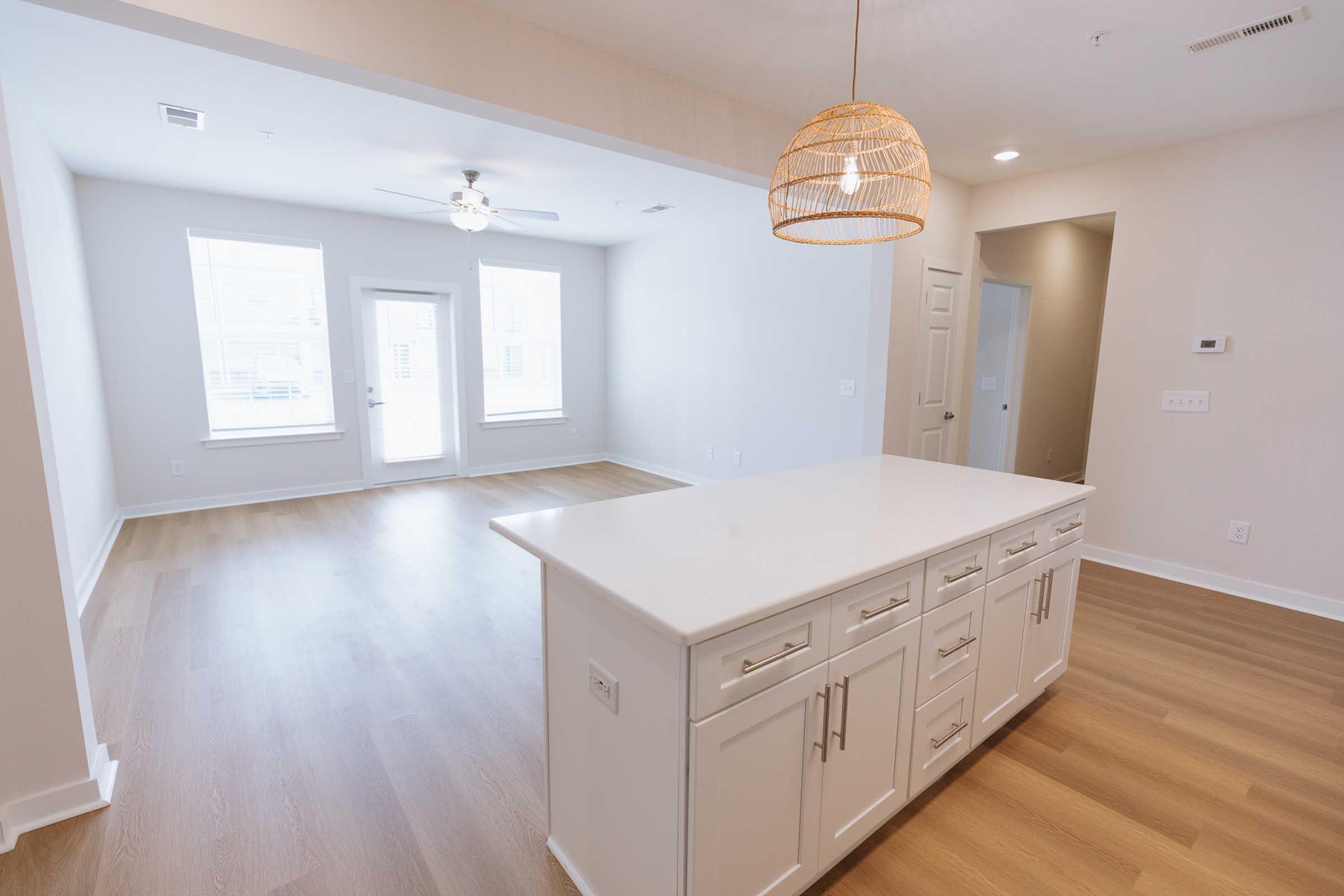 A bright, spacious living area featuring light-colored walls and wooden flooring. The room includes a central kitchen island with cabinets, a ceiling fan, and a doorway leading to an outdoor patio. Large windows provide natural light, creating an inviting atmosphere.