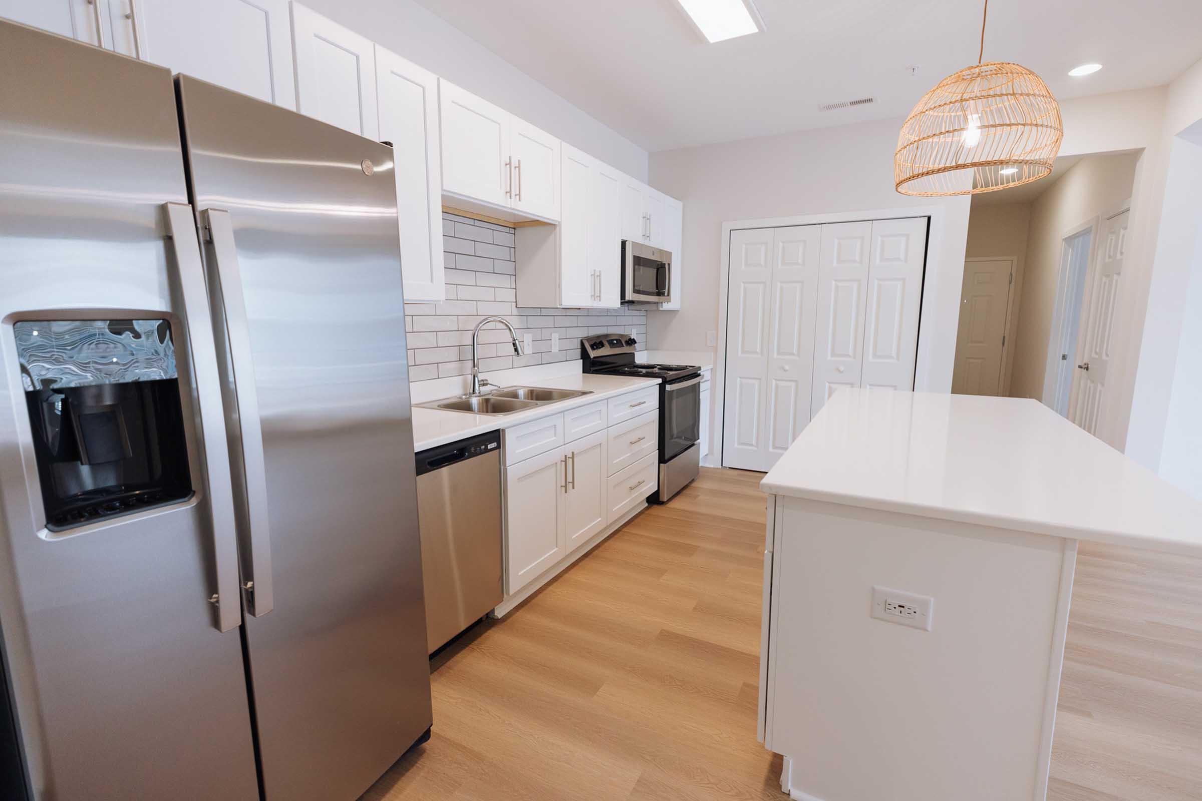 Modern kitchen featuring stainless steel appliances, including a fridge with a water dispenser, a stove, and a microwave. The cabinetry is white with a light grey backsplash, and there’s a large island with a white countertop. A pendant light hangs above the island in a warm wood tone.