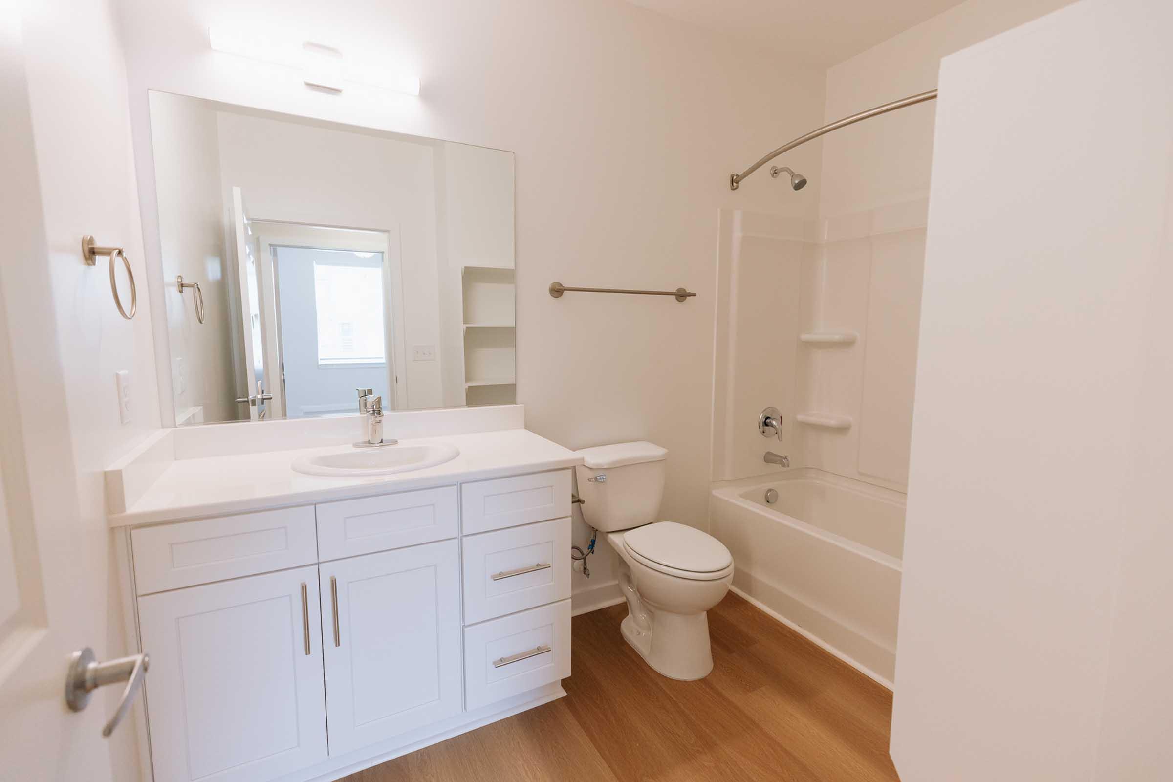 Bright and modern bathroom featuring a white vanity with a sink, large mirror, and a walk-in shower/bathtub combination. The walls are painted light, and the flooring is wooden. The space includes a toilet, towel bar, and shelving for storage. Natural light enters from a nearby window.