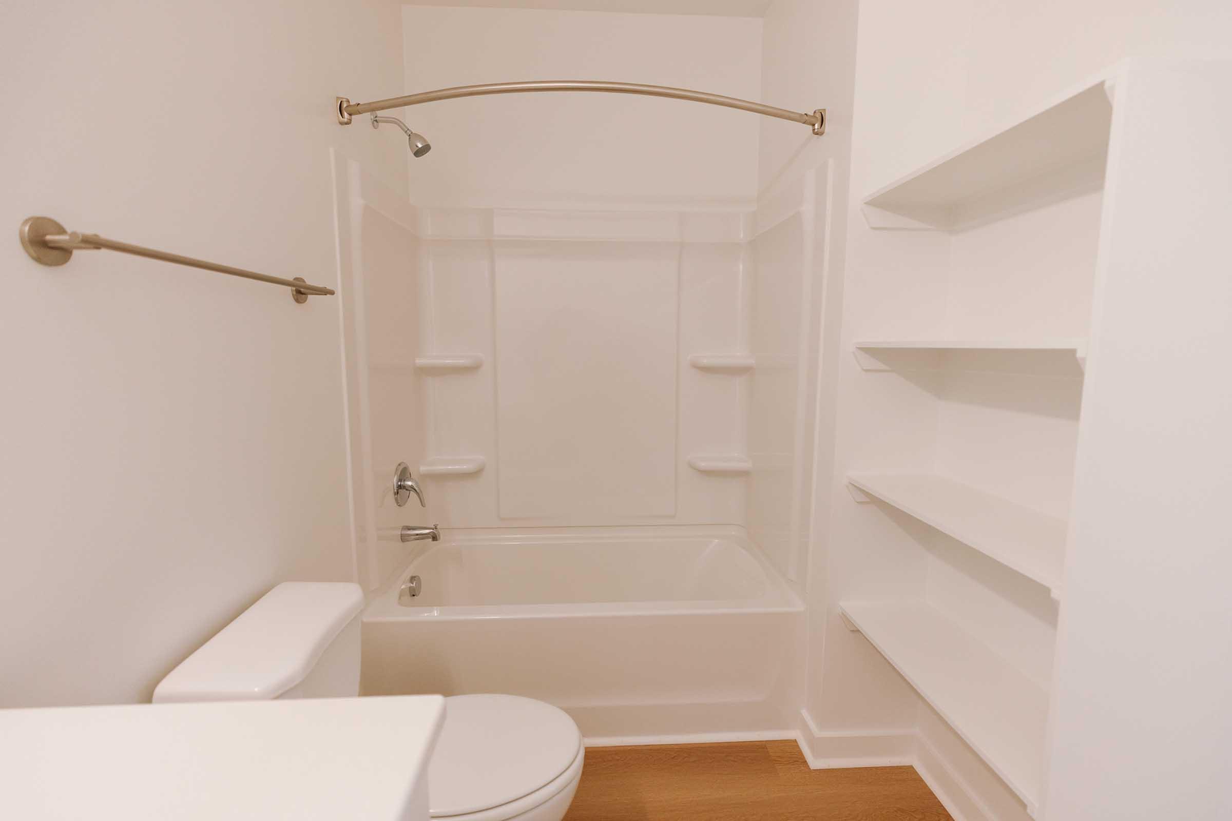 A clean, modern bathroom featuring a bathtub with a showerhead, a toilet, and two wall-mounted shelves. The walls are white, and the flooring is light wood. The design is minimalistic and bright, with a curved shower rod above the tub.