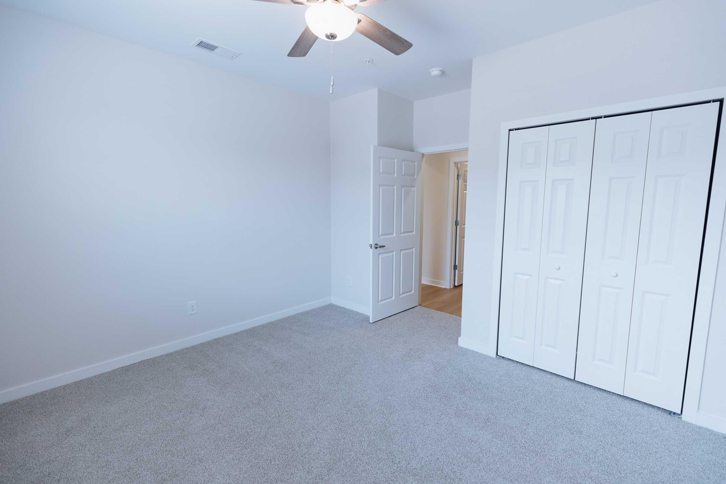 A spacious, empty room with light-colored walls and carpeted flooring. There is a ceiling fan and a door leading to another area. Two double sliding closet doors are visible along one wall. The room is well-lit, creating a bright and airy atmosphere.