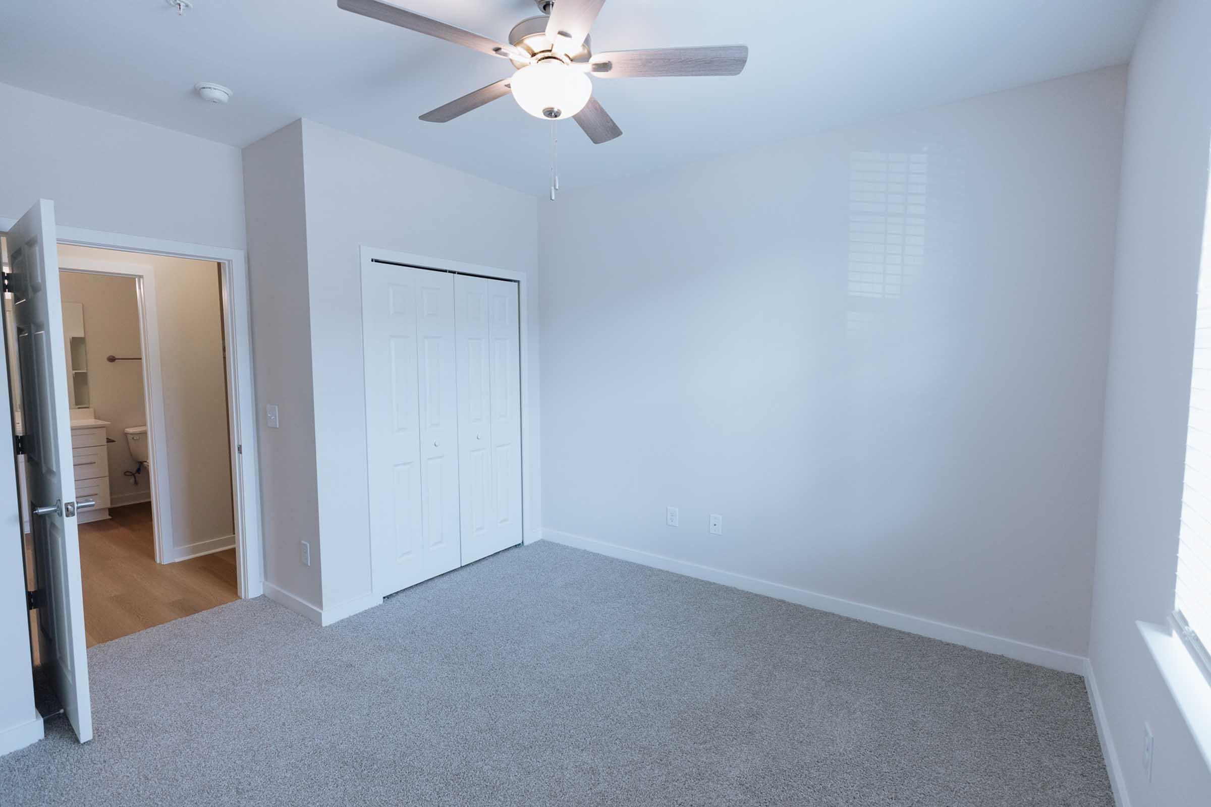 A spacious, well-lit bedroom with light-colored walls and soft gray carpet. It features a ceiling fan, a closet with sliding doors, and a doorway leading to a bathroom. Natural light enters through the window, enhancing the room's inviting atmosphere.